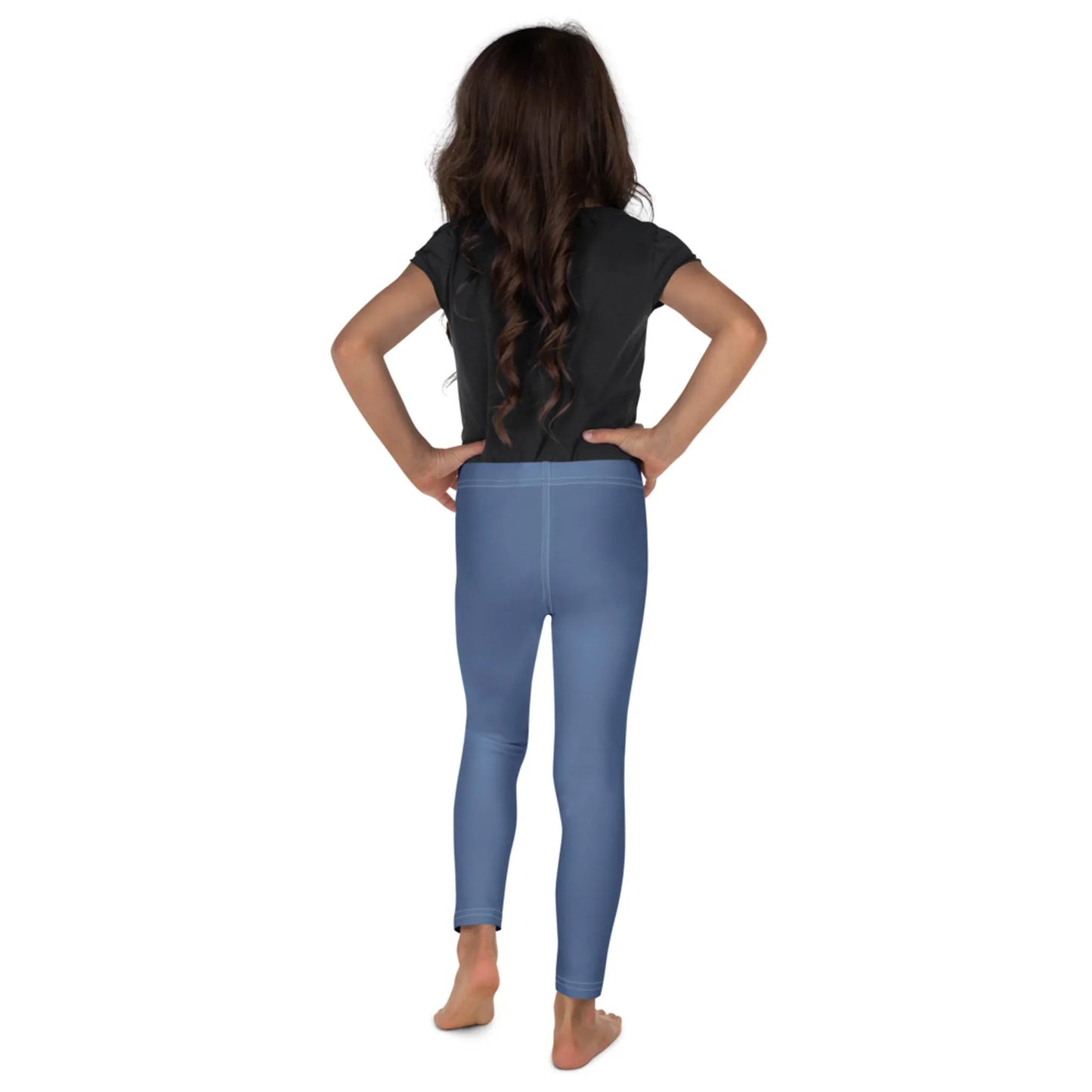 A young girl with long brown hair stands barefoot, facing away with hands on hips. She wears a black Mambí t-shirt and light blue leggings against a plain white background.