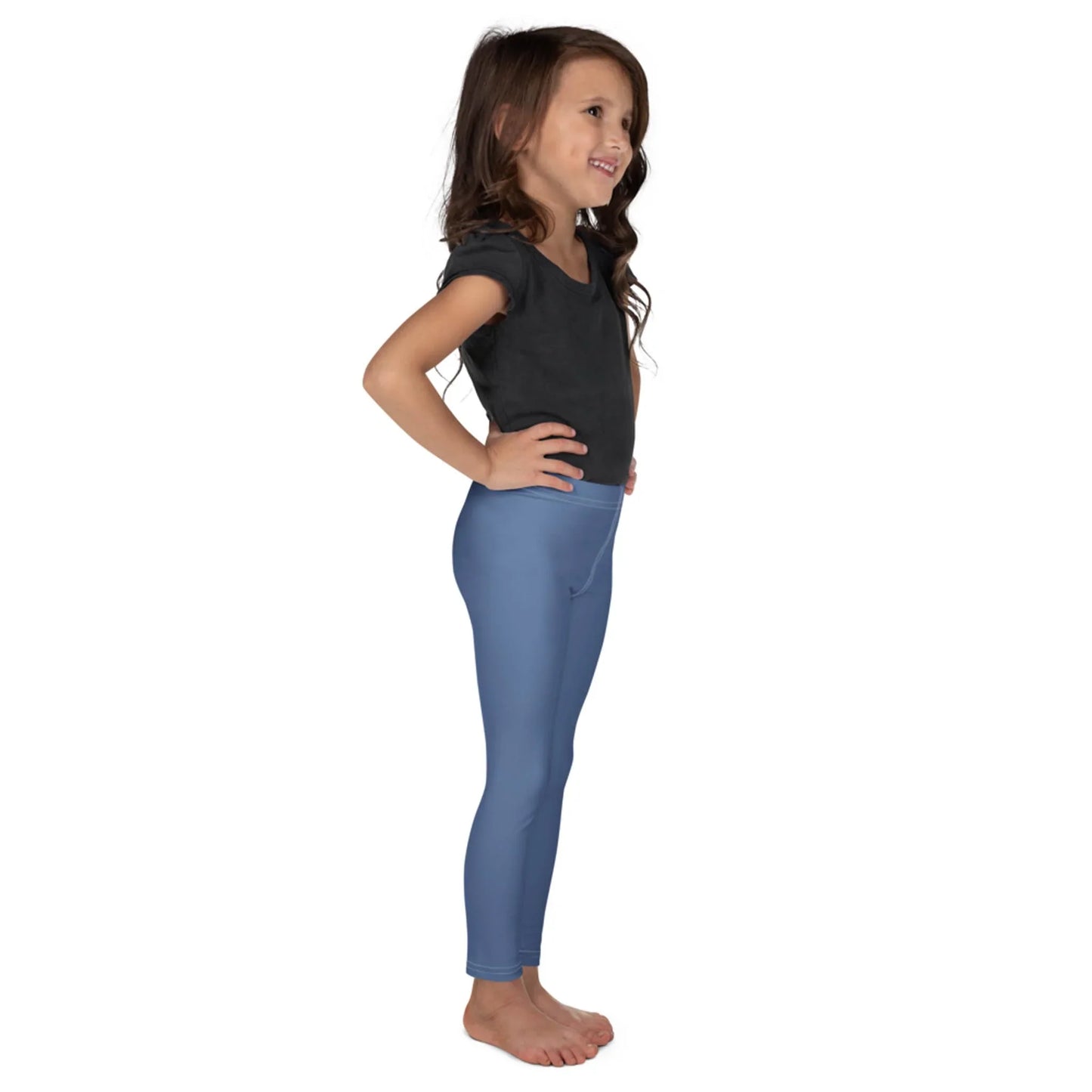 Young girl standing in a side view, smiling, with long brown hair, wearing a black Mambí t-shirt and light blue leggings, barefoot, against a plain white background.