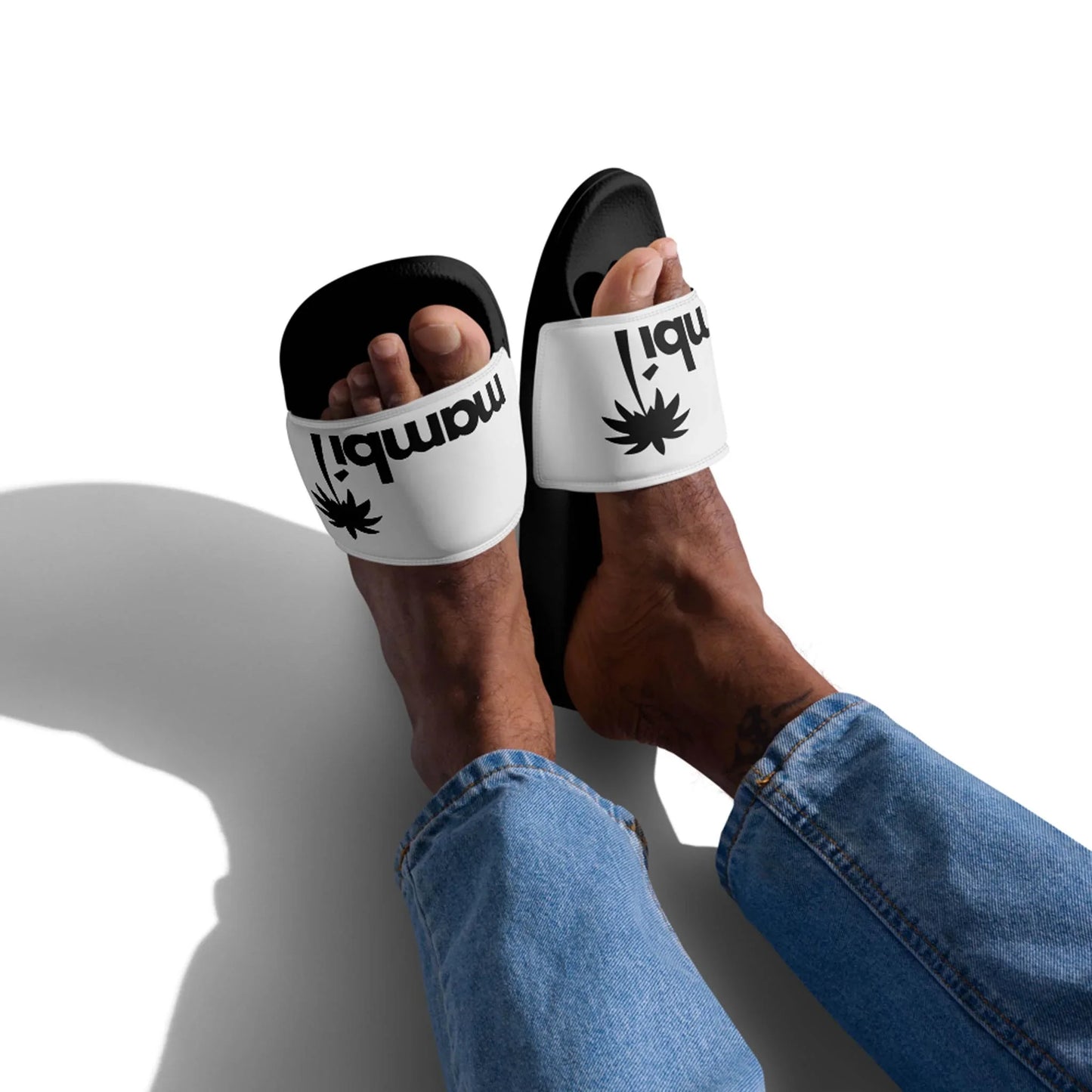 A person wearing blue jeans and Printful Malibu Chancleta Slides for men in Black/Whisper, featuring cushioned white straps with a black graphic and the word Mombi! printed upside down. Plain white background with a visible shadow.