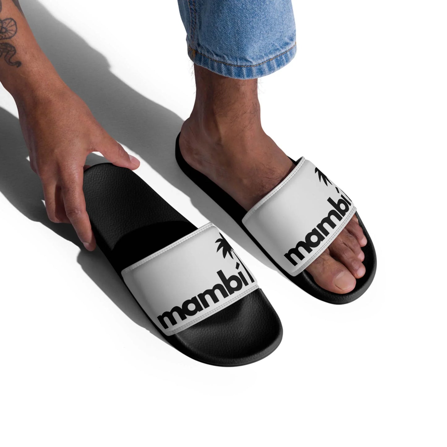 A person in blue jeans is slipping on Printful Malibu Chancleta Slides for men in Black/Whisper, featuring mambi and a palm tree graphic on the cushioned upper strap. Only their lower legs and hand are visible against a white background.