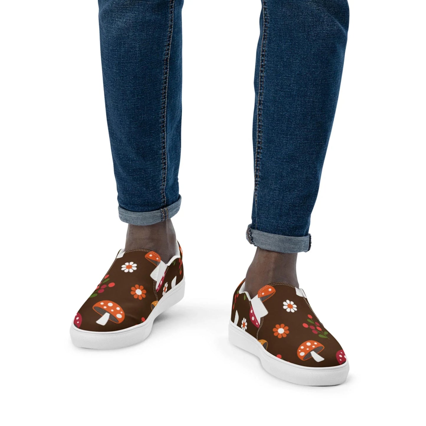A person stands against a brown background with a vibrant mushroom and flower pattern, wearing dark blue rolled-up jeans and Mario Handmade Slip-On Canvas Shoes for men by Printful, making a playful fashion statement.