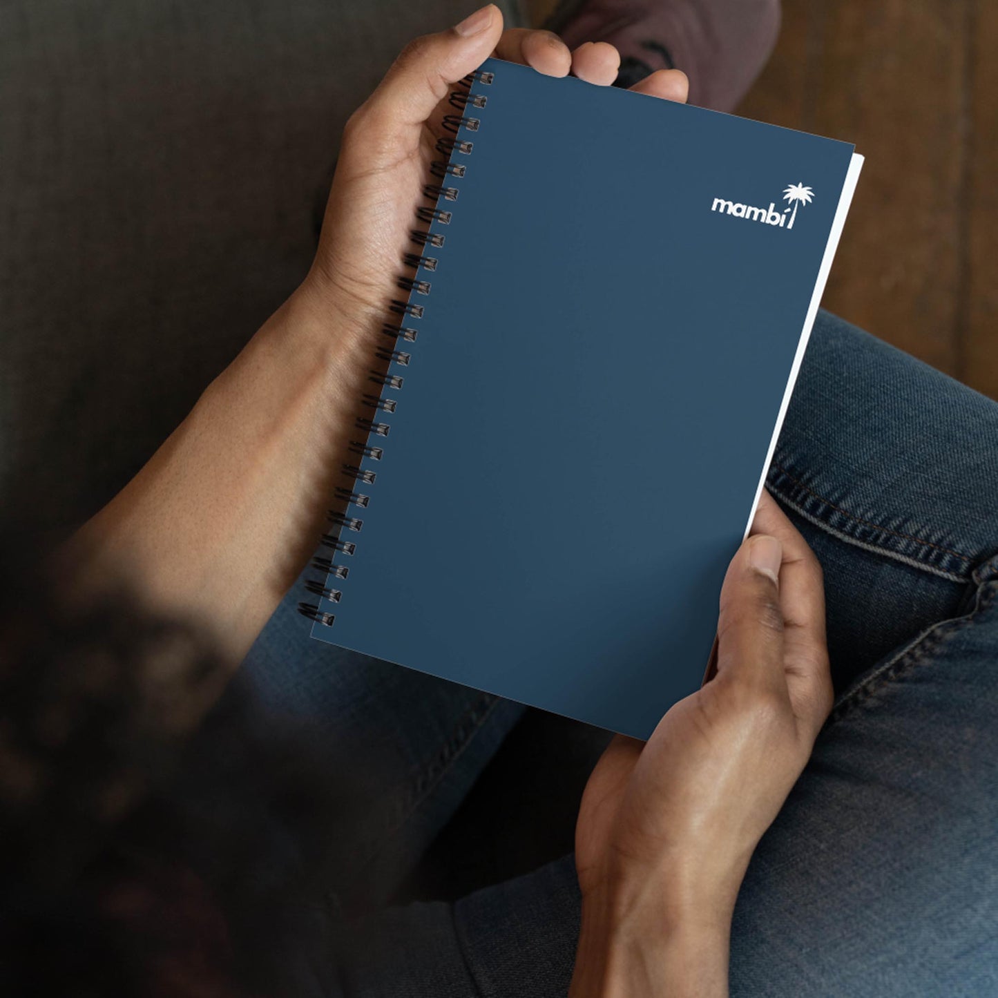 A person sits cross-legged, holding a custom wire-bound notebook with a blue cover, a white mambi logo, and a palm tree in the top right corner.