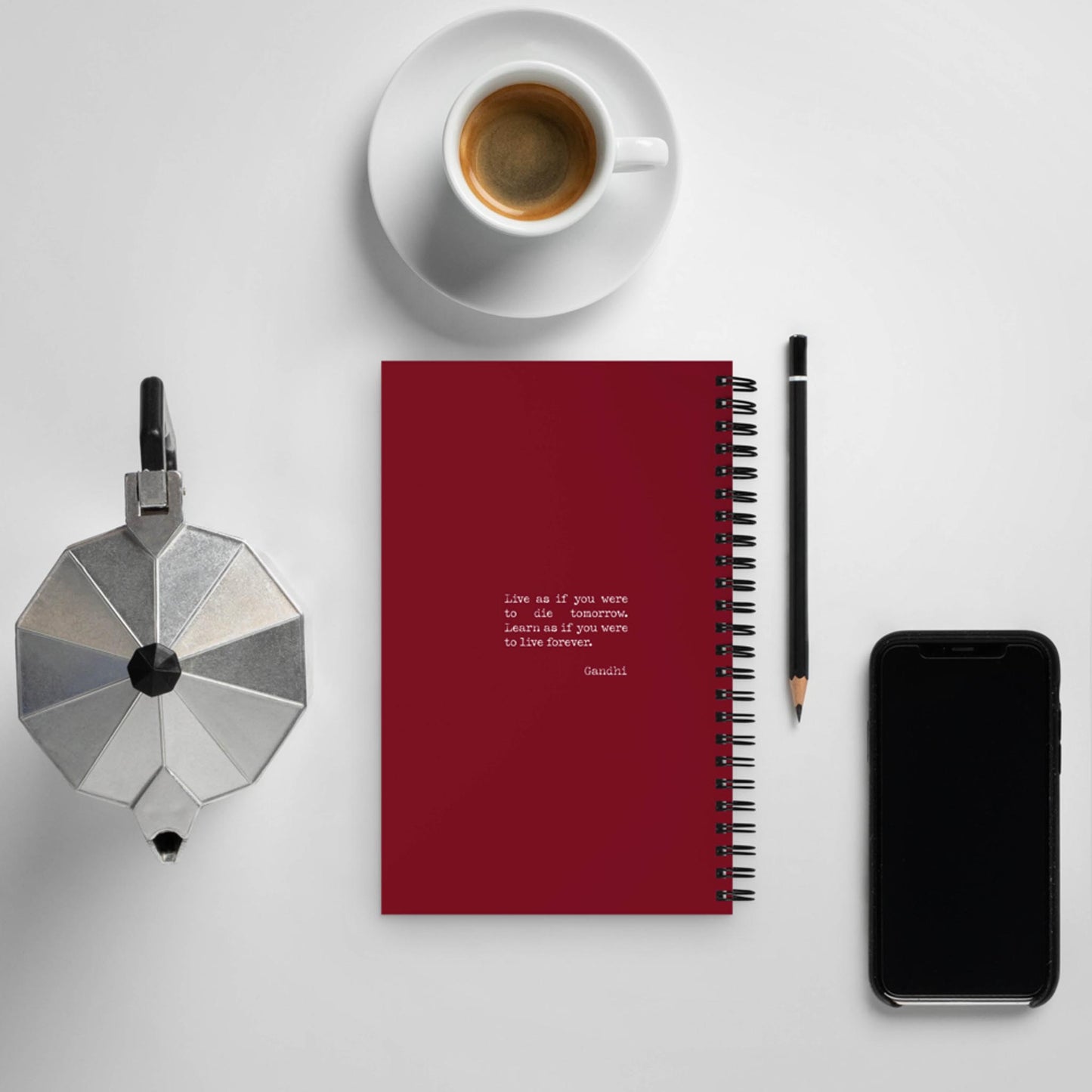 A flat lay of a red spiral notebook, a pencil, a cup of coffee on a saucer, a moka pot, and a smartphone on a white surface. The custom wire-bound notebook features an inspiring quote printed on the cover.