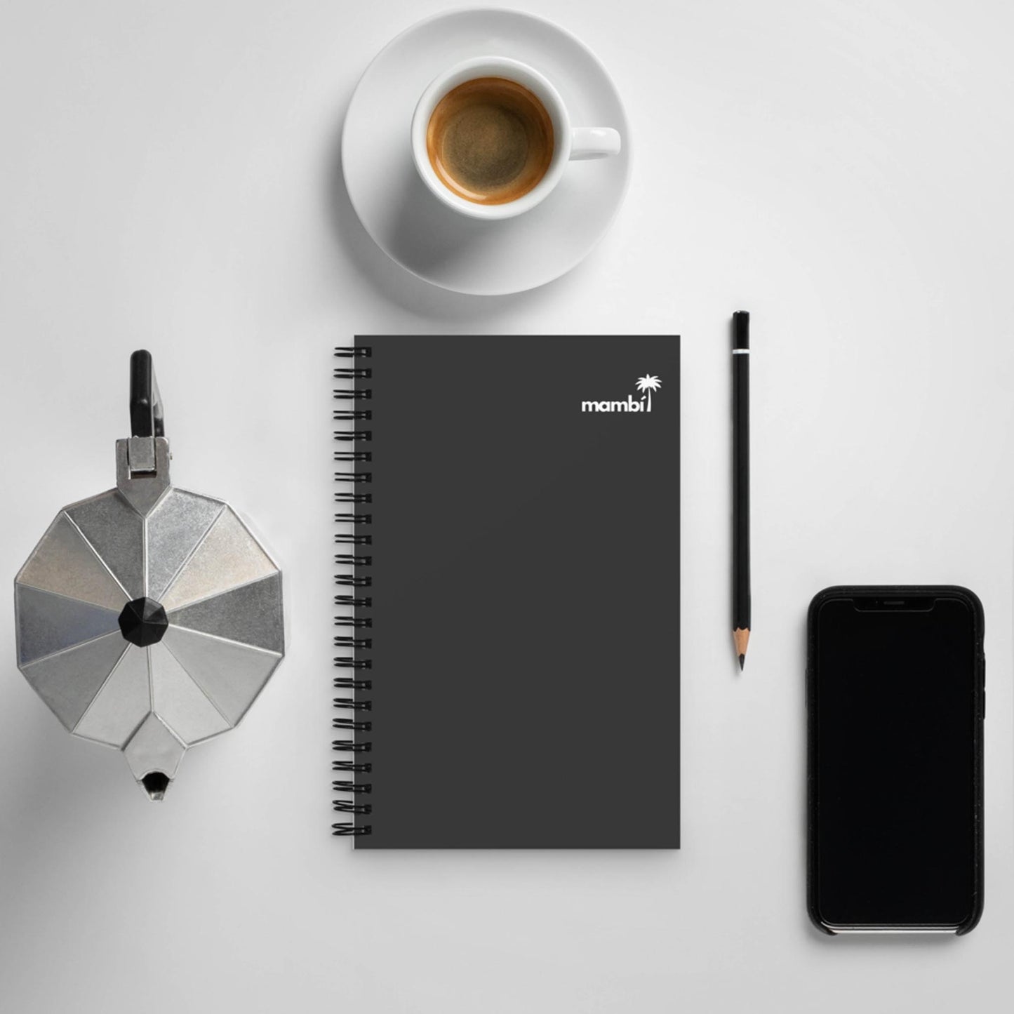 A custom wire-bound notebook, black pencil, smartphone, cup of coffee, and a stovetop espresso maker are arranged neatly on a white surface.