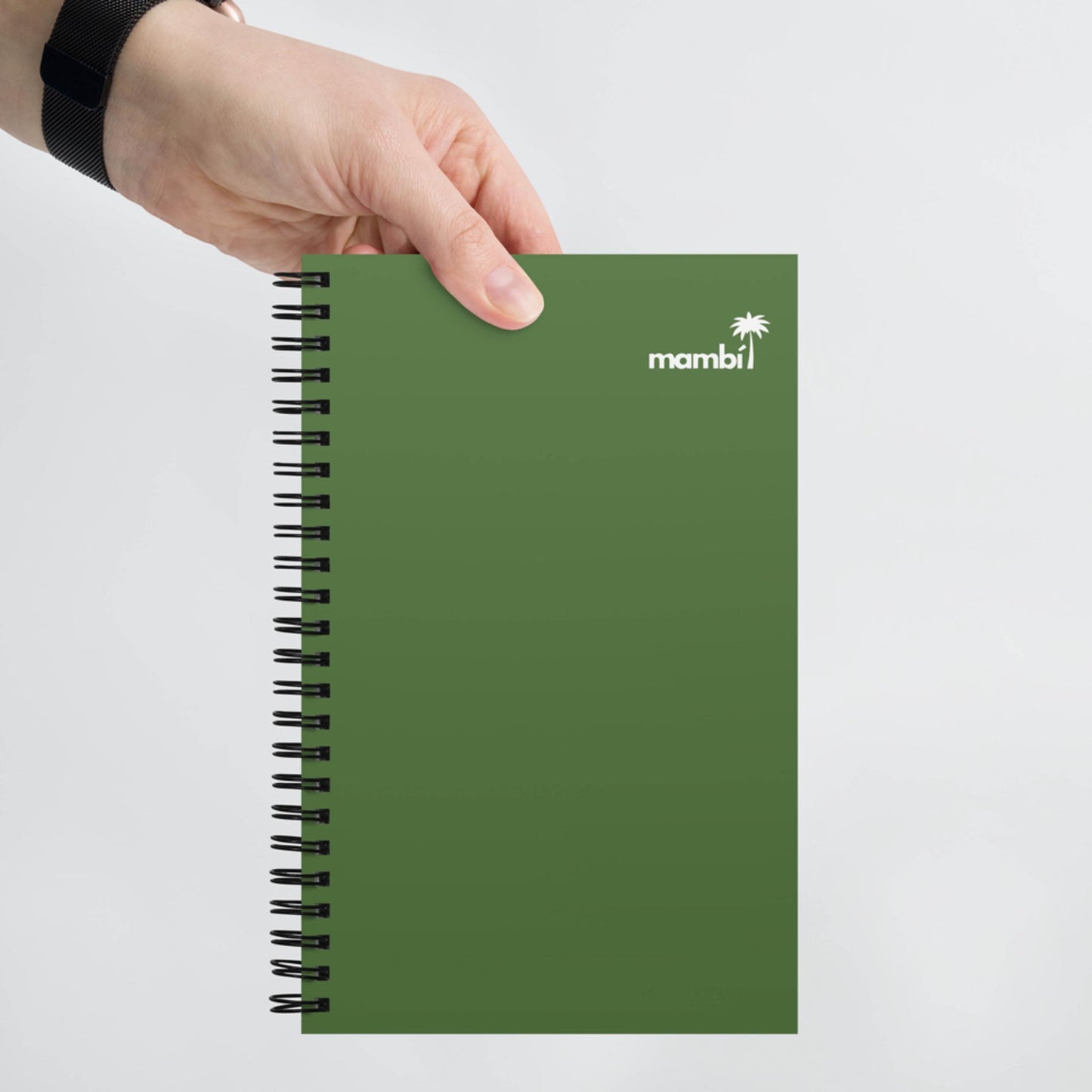 A hand holding a green, spiral notebook with mambi and a small palm tree logo printed in white on the top right corner of the cover.