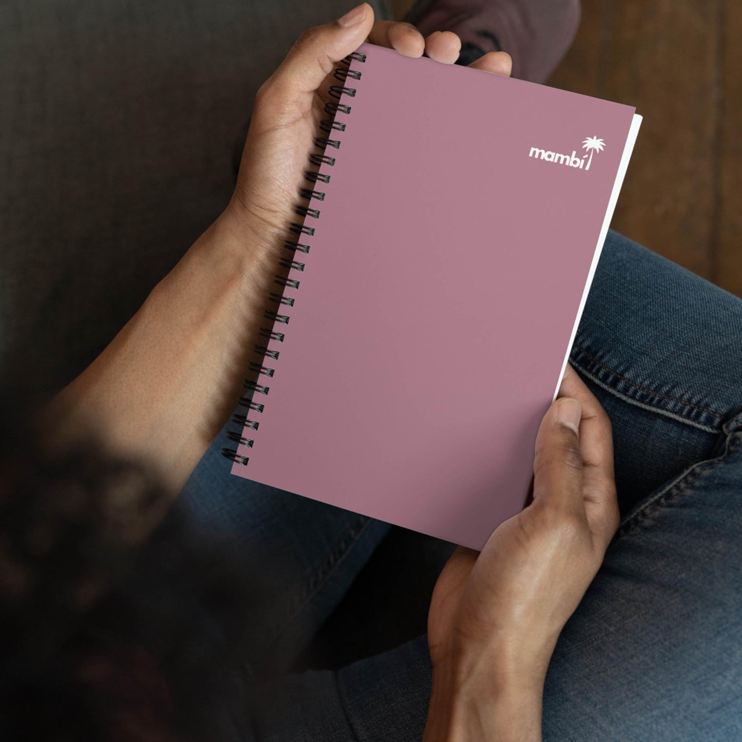A person holds a closed, pink custom wire-bound notebook with the word mambi and a palm tree logo on the cover while sitting cross-legged.