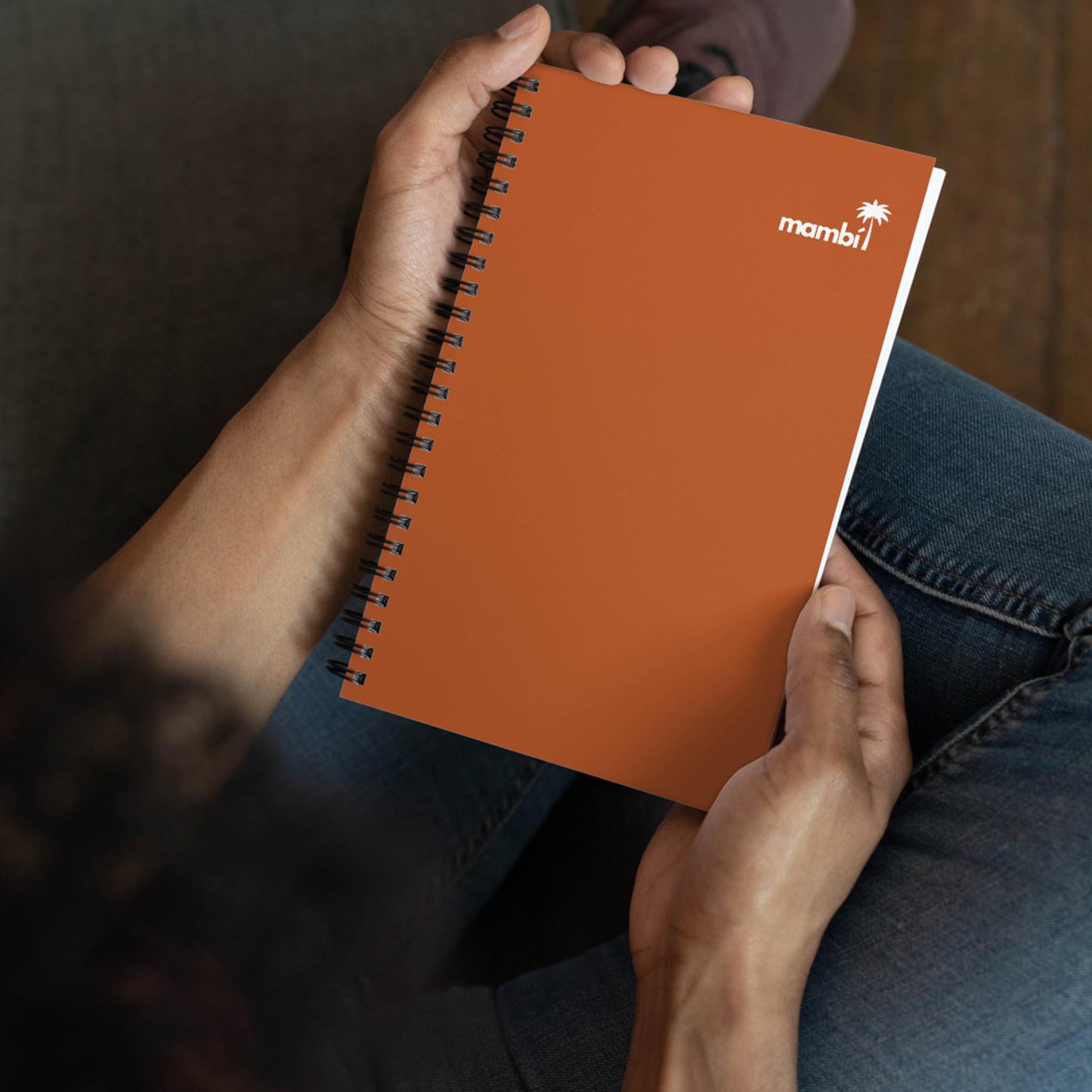 A person sits cross-legged, holding an orange wire-bound notebook with the word mambi and a small palm tree icon printed in white on the cover.