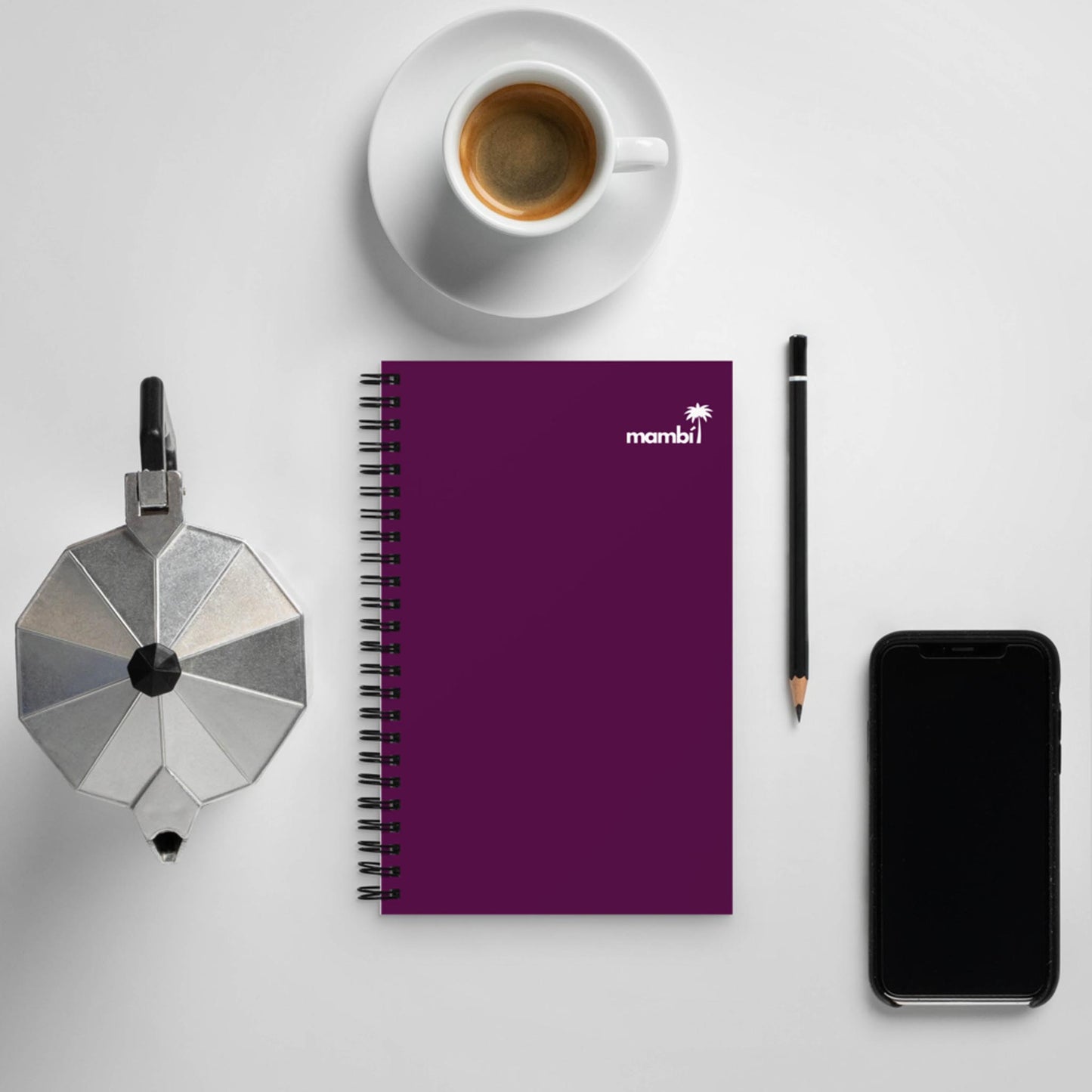 A flat lay image showing a white cup of coffee, a silver stovetop espresso maker, a purple wire-bound notebook, a black pencil, and a black smartphone on a white surface.