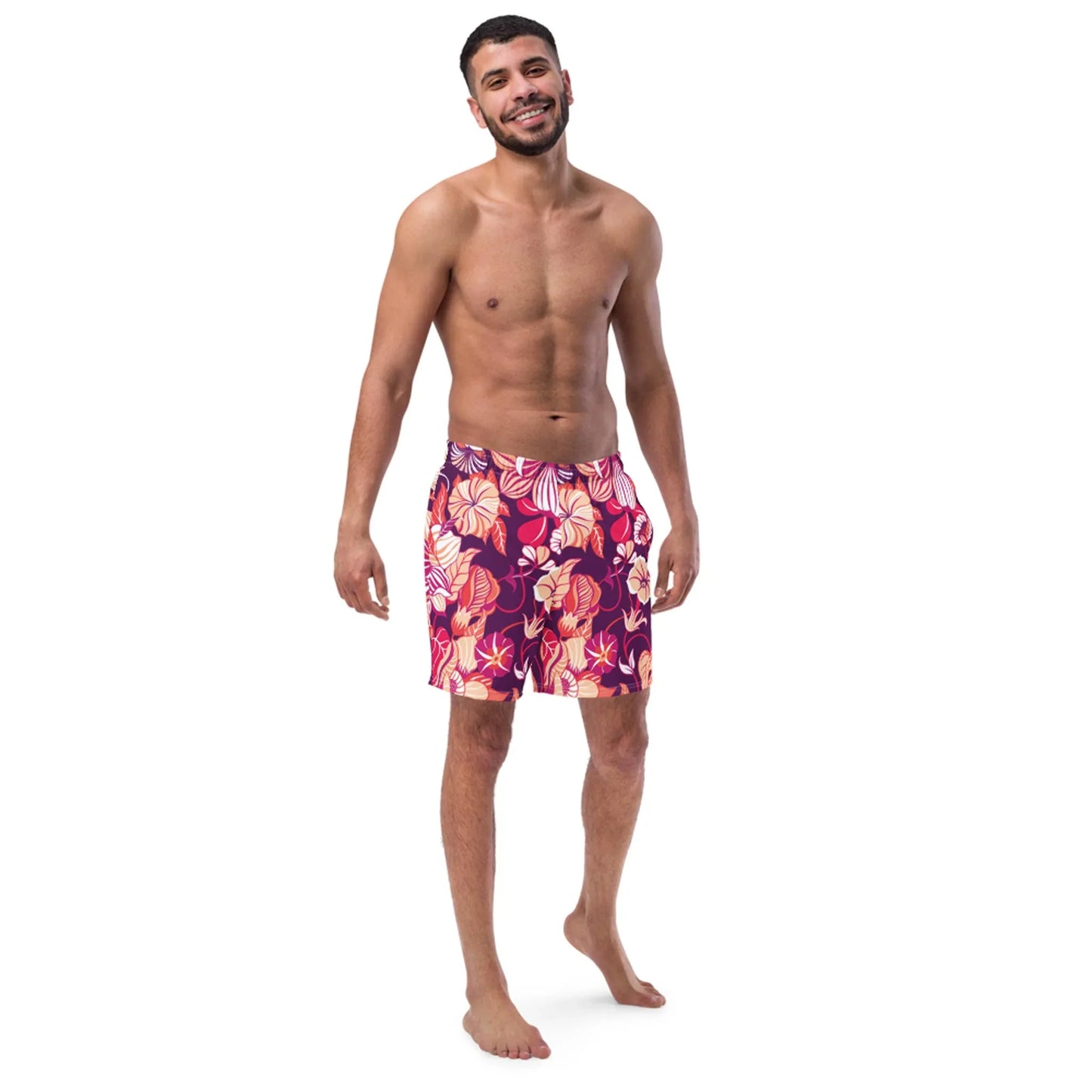 A smiling, barefoot man with short dark hair and a beard stands against a white background wearing Printful Mauricio Mens Swim Trunks in vibrant red, orange, and purple. Features a comfortable anti-chafe inner liner.