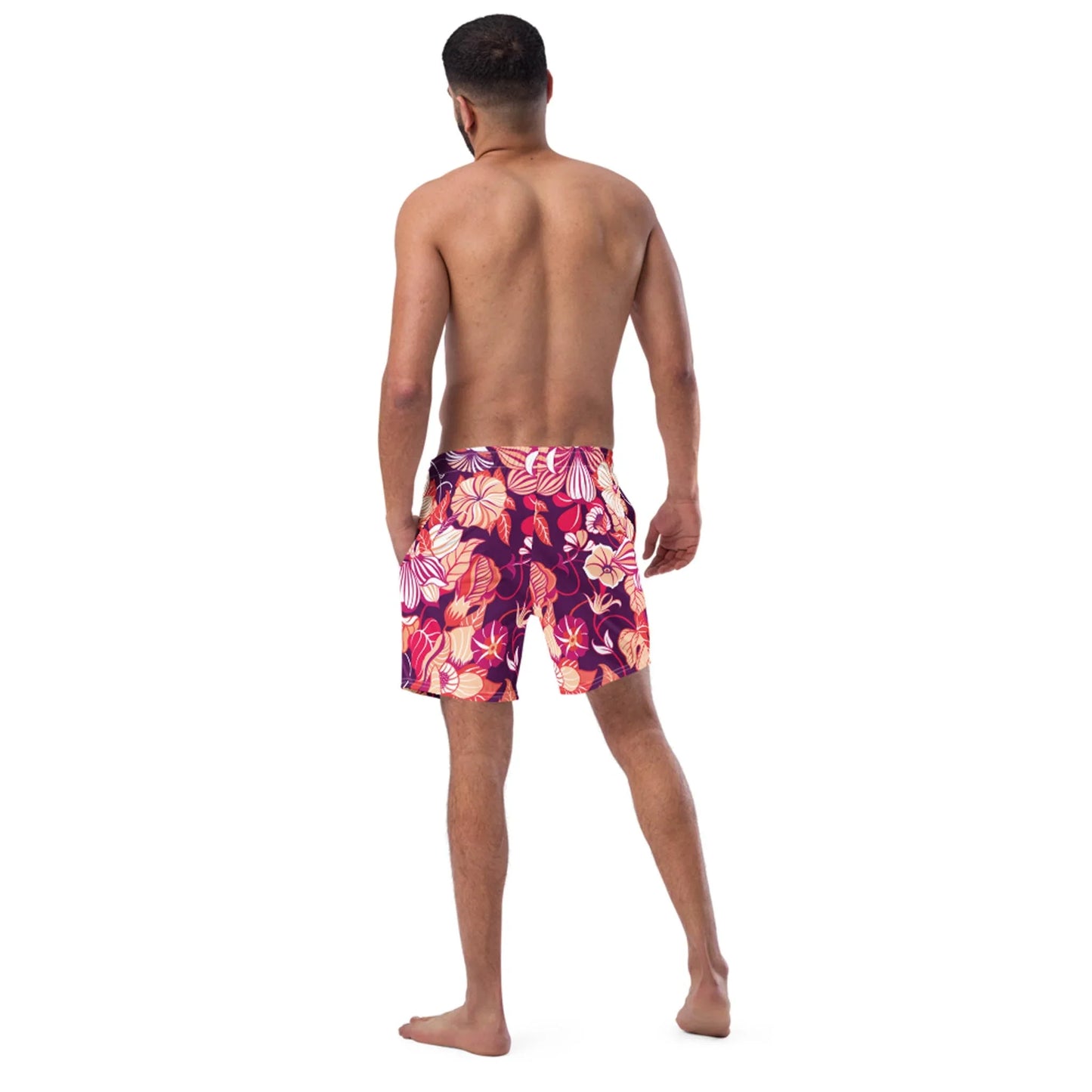 A man stands barefoot with his back to the camera, wearing Printful Mauricio Swim Trunks for men featuring a colorful floral pattern on soft microfiber, set against a plain white background.
