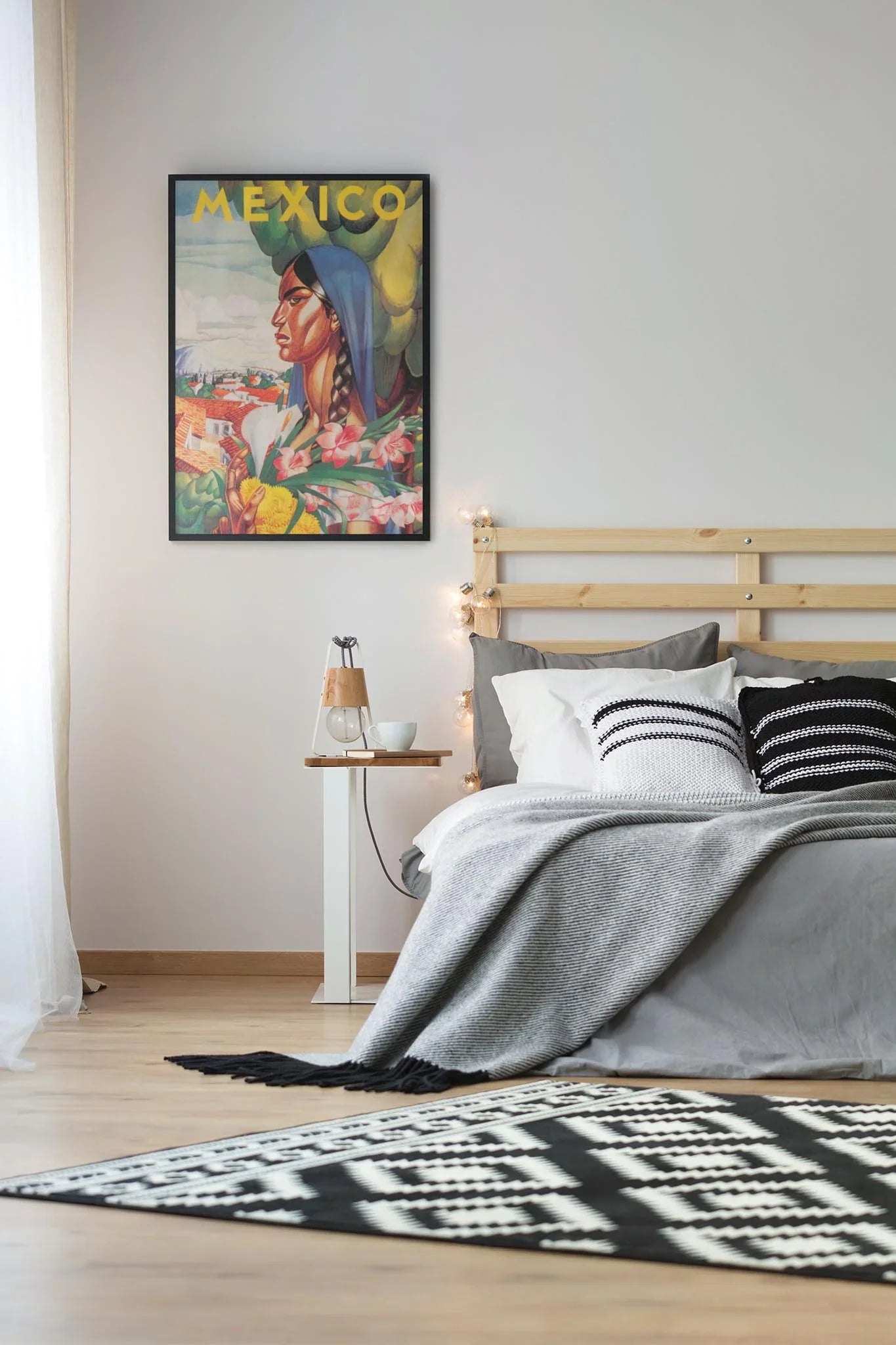 A cozy bedroom features a wooden bed, gray and black bedding, geometric rug, bedside table with a lamp, string lights, and the Mexico Reprinted Vintage Poster by Printful (24x36, premium luster paper) above the bed for added style.