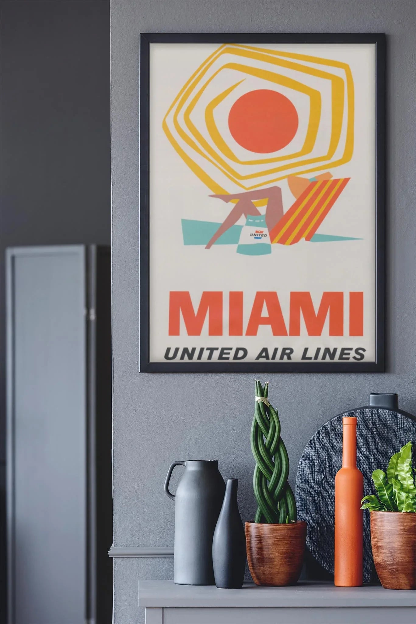 A framed vintage United Air Lines Miami travel poster featuring a beach scene and a touch of Mambí style hangs on a gray wall above a shelf adorned with vases, a potted plant, and a round black tray.