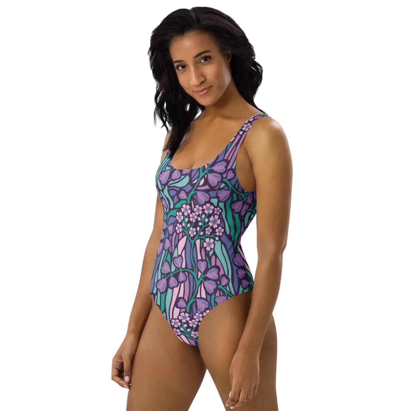 A woman with shoulder-length black hair wears the Micaela One-Piece Swimsuit by Printful in a chic purple and green floral design, standing against a white background and smiling slightly at the camera.