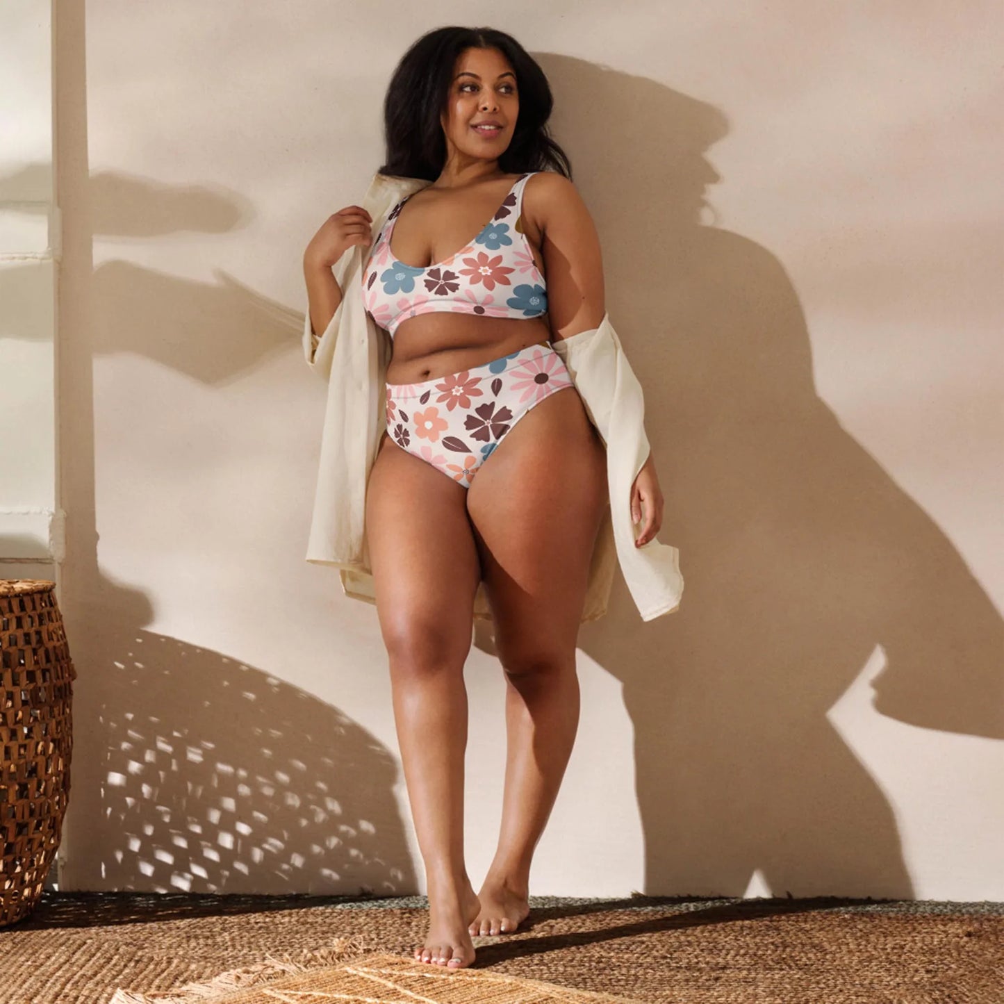 A woman stands barefoot indoors on a woven rug, wearing a floral bikini and an open Mambí light-colored shirt. She smiles and poses confidently against a neutral wall with sunlight casting soft shadows.