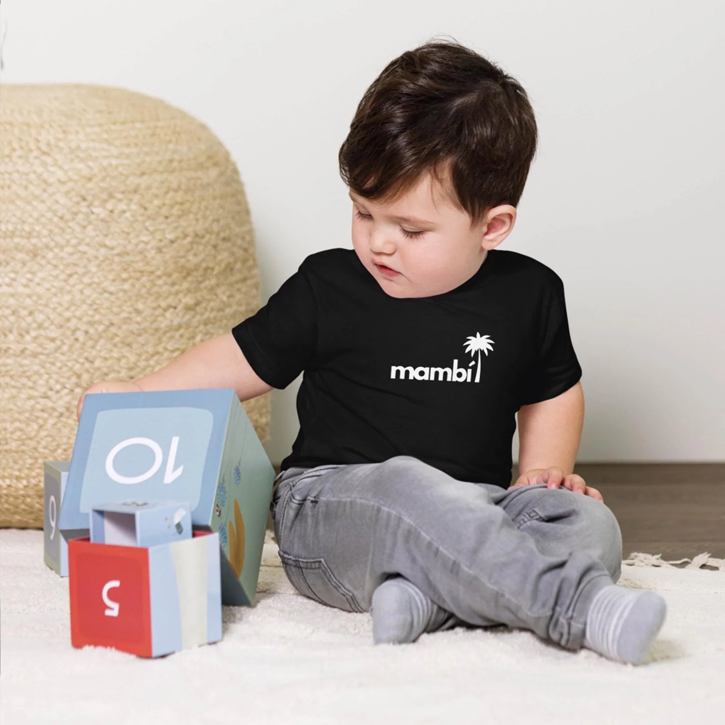 A young child with short dark hair sits on the floor, wearing a black Mambí t-shirt and gray pants, playing with colorful stacking blocks. A woven basket is in the background.