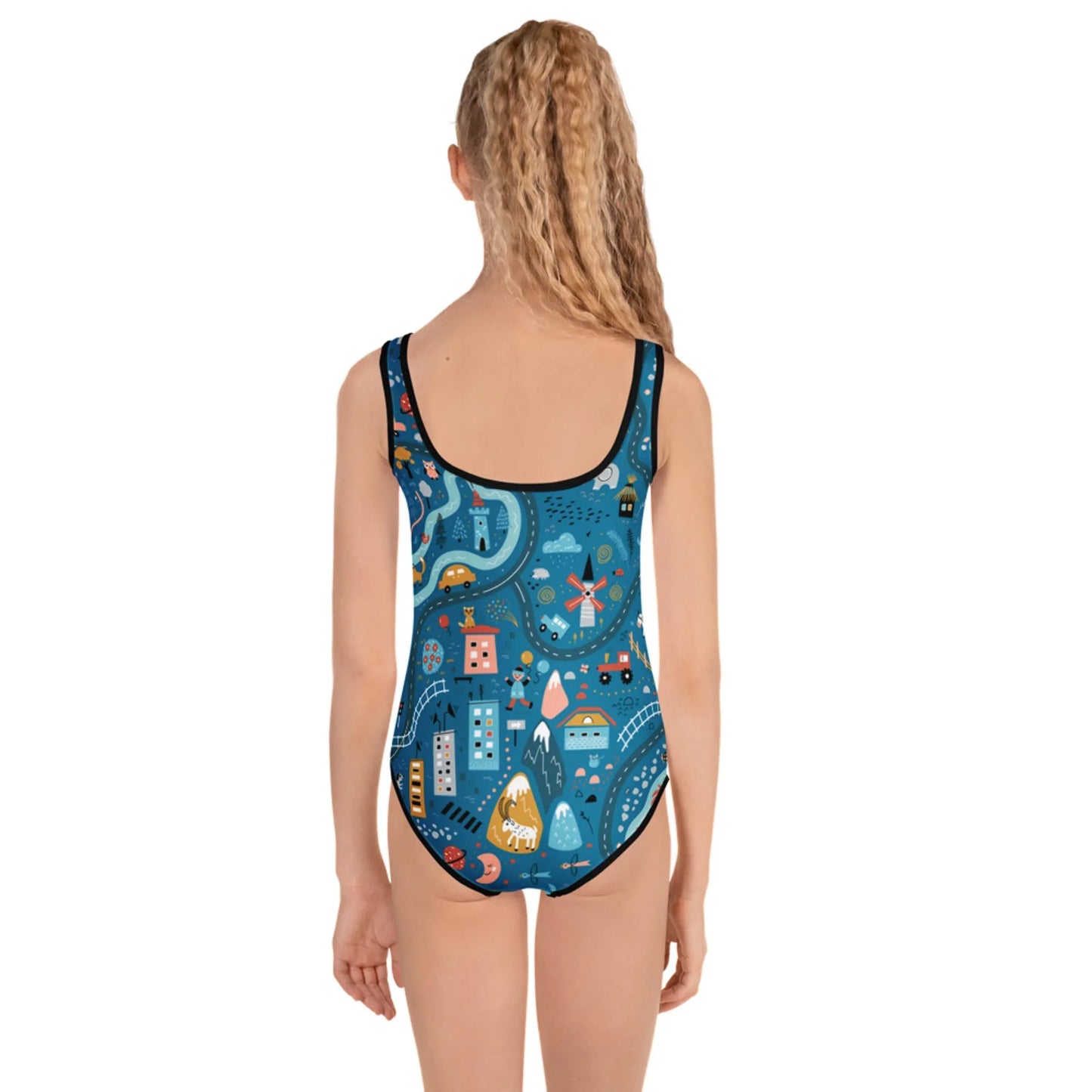 A girl with long, wavy blonde hair is shown from the back, wearing a Mambí one-piece swimsuit featuring a colorful, whimsical cityscape pattern on a blue background.