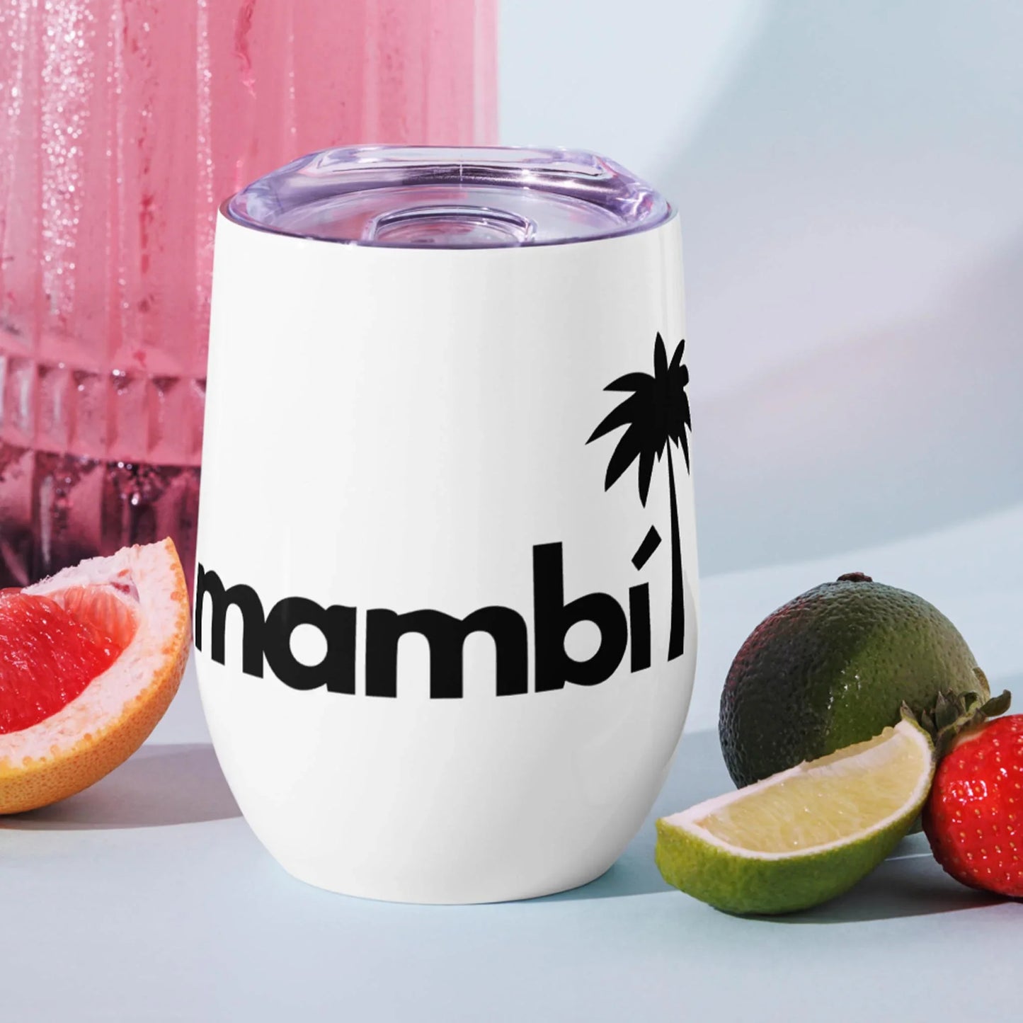 A white tumbler with a transparent lid, featuring the black text Mambí and a palm tree graphic. Surrounded by sliced grapefruit, lime, a strawberry, and a green fruit, with a pink drink in the background.