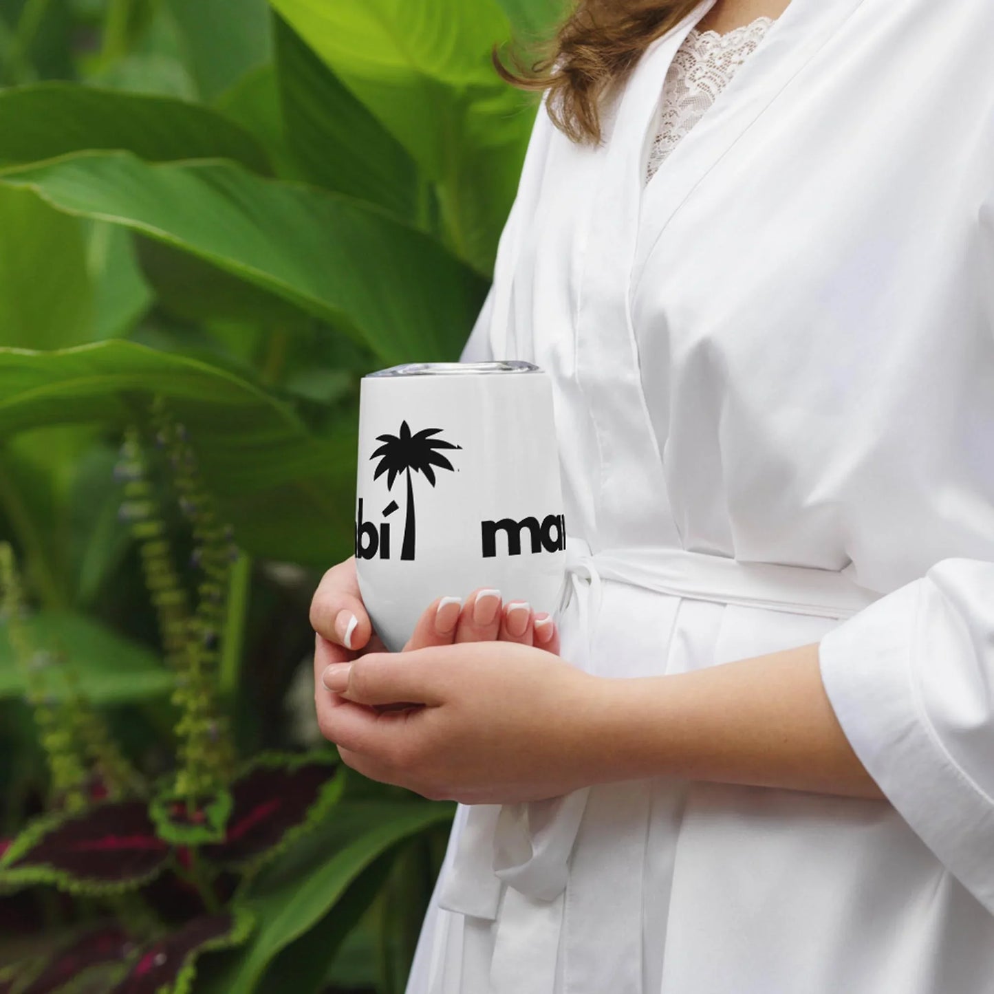 A person in a white robe holds a white cup featuring a black palm tree graphic and partial Mambí text, standing in front of lush green plants.