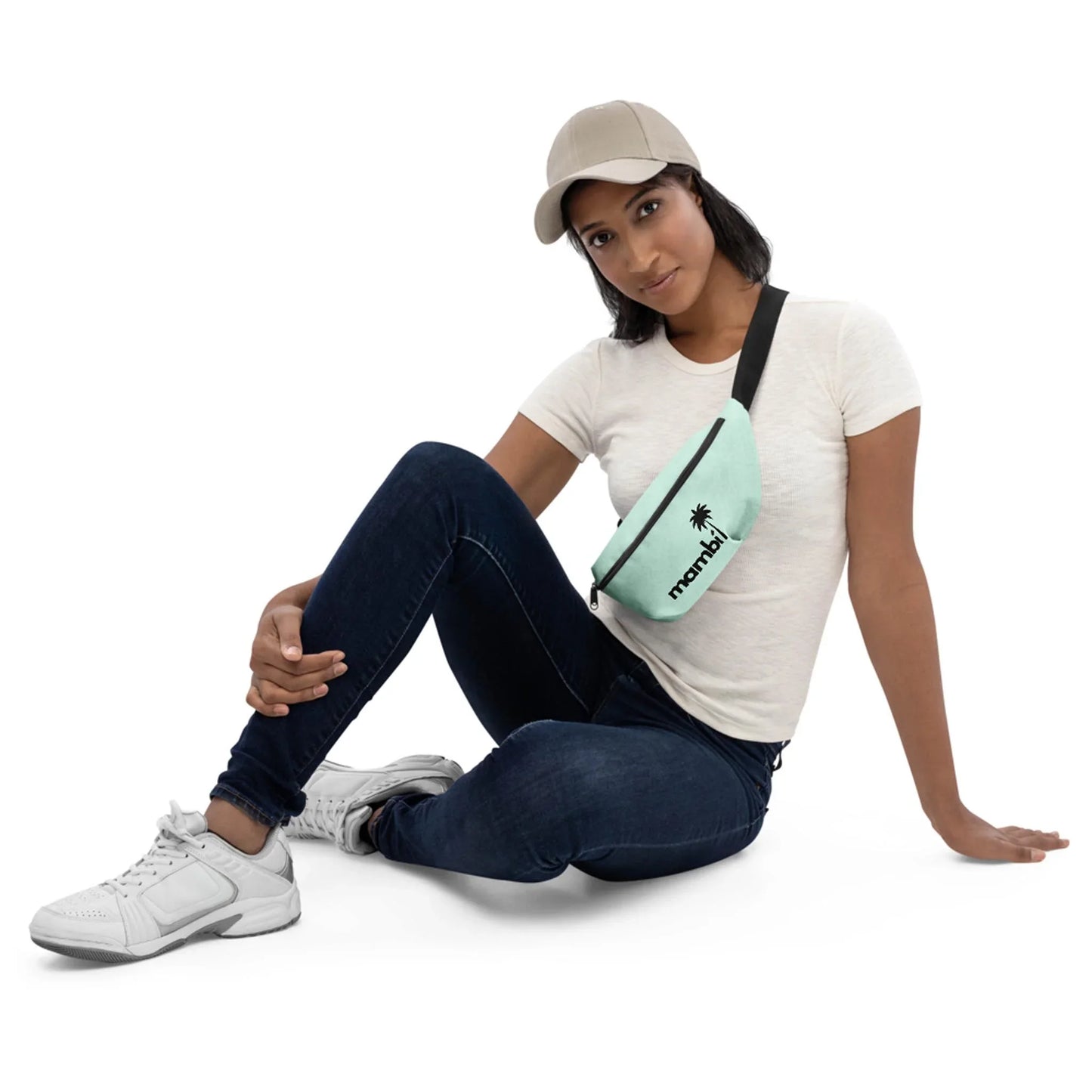 Woman in a beige cap, white t-shirt, and jeans sits on the floor with one knee up, wearing white sneakers and a light blue Mambí crossbody bag that says momiji with a palm tree icon. The background is white.