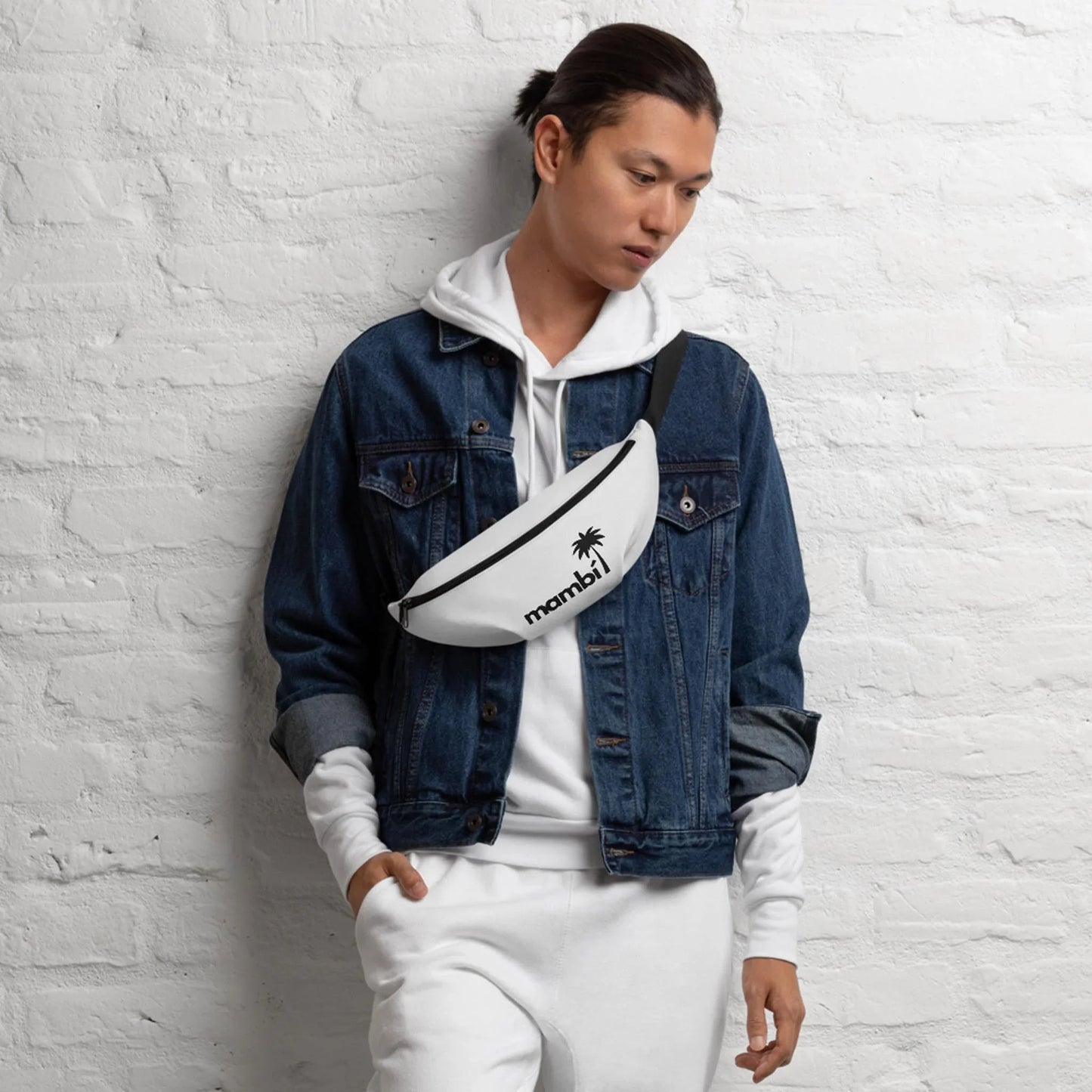 A person in a white hoodie and sweatpants, a denim jacket, and a white crossbody bag with the Mambí palm tree logo stands against a white brick wall, looking down with one hand in their pocket.