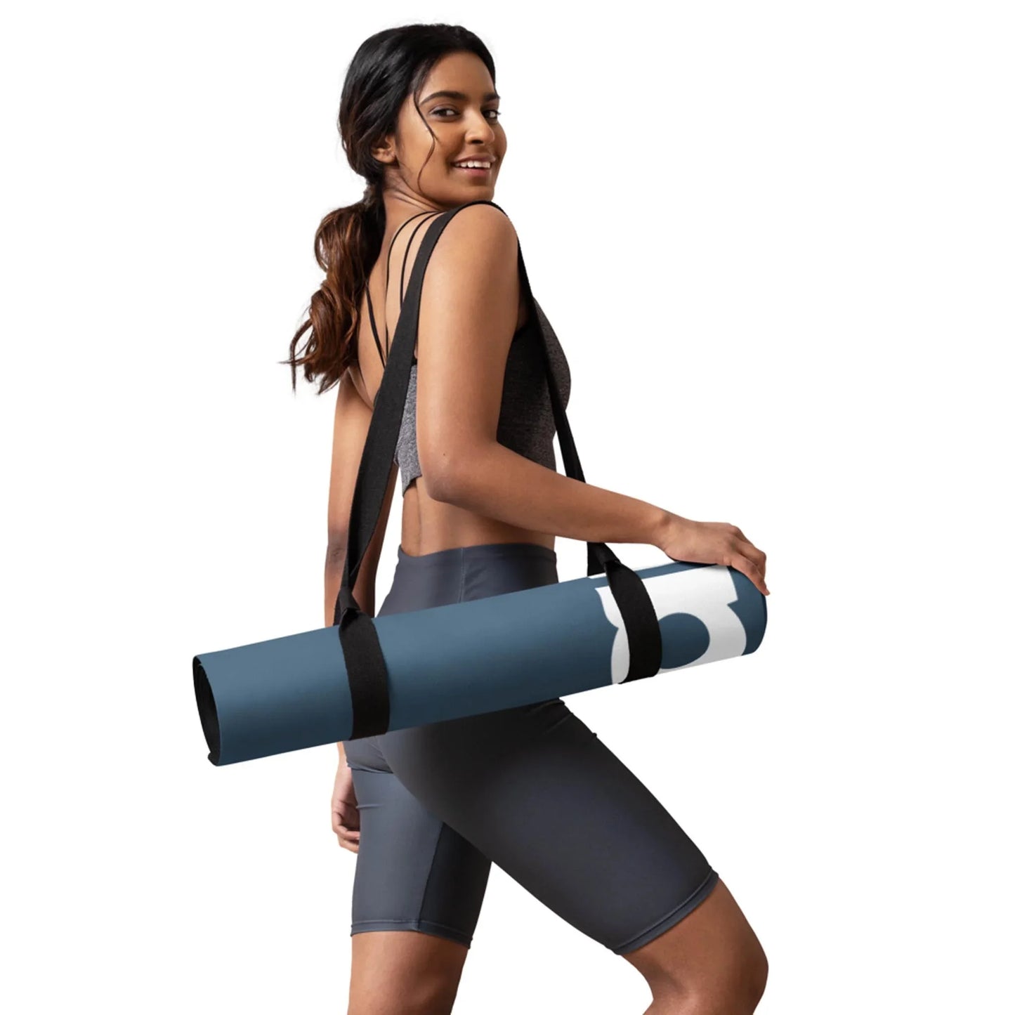 A woman in athletic wear smiles while holding a rolled-up blue Mambí yoga mat with a black strap over her shoulder, standing against a white background.