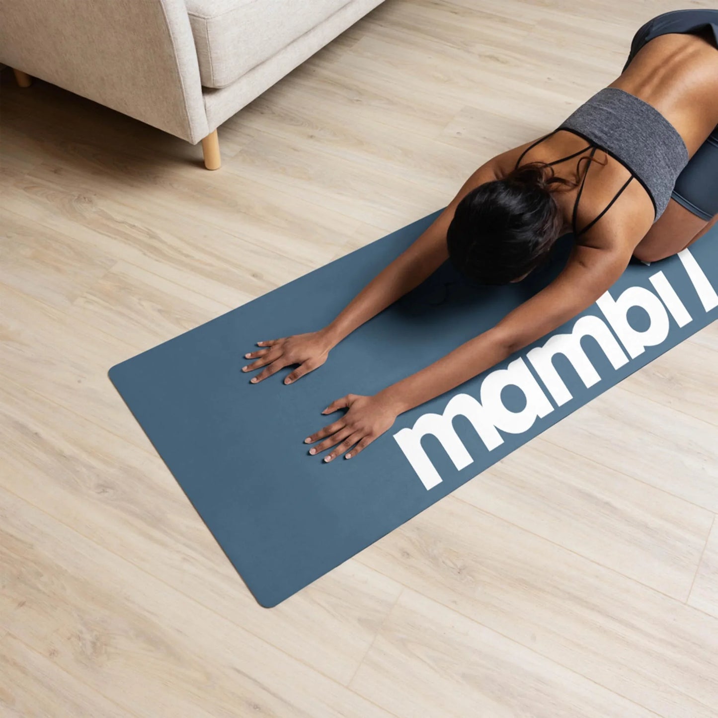 A woman in athletic wear is doing a childs pose yoga stretch on a blue mat with the word Mambí visible, on a light wood floor next to a beige sofa.