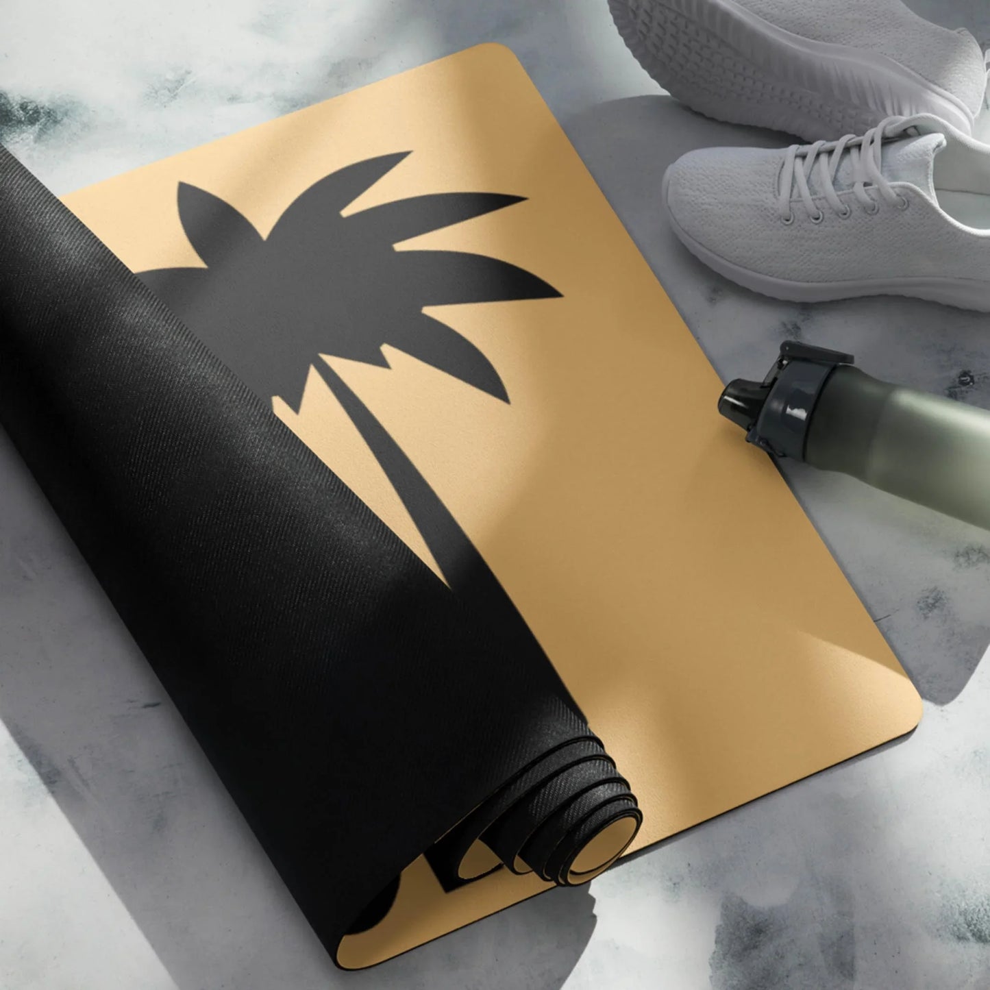 A rolled-up black Mambí yoga mat partially covers a tan mat with a black palm tree design. White athletic shoes and a green water bottle are nearby on a light marble floor.