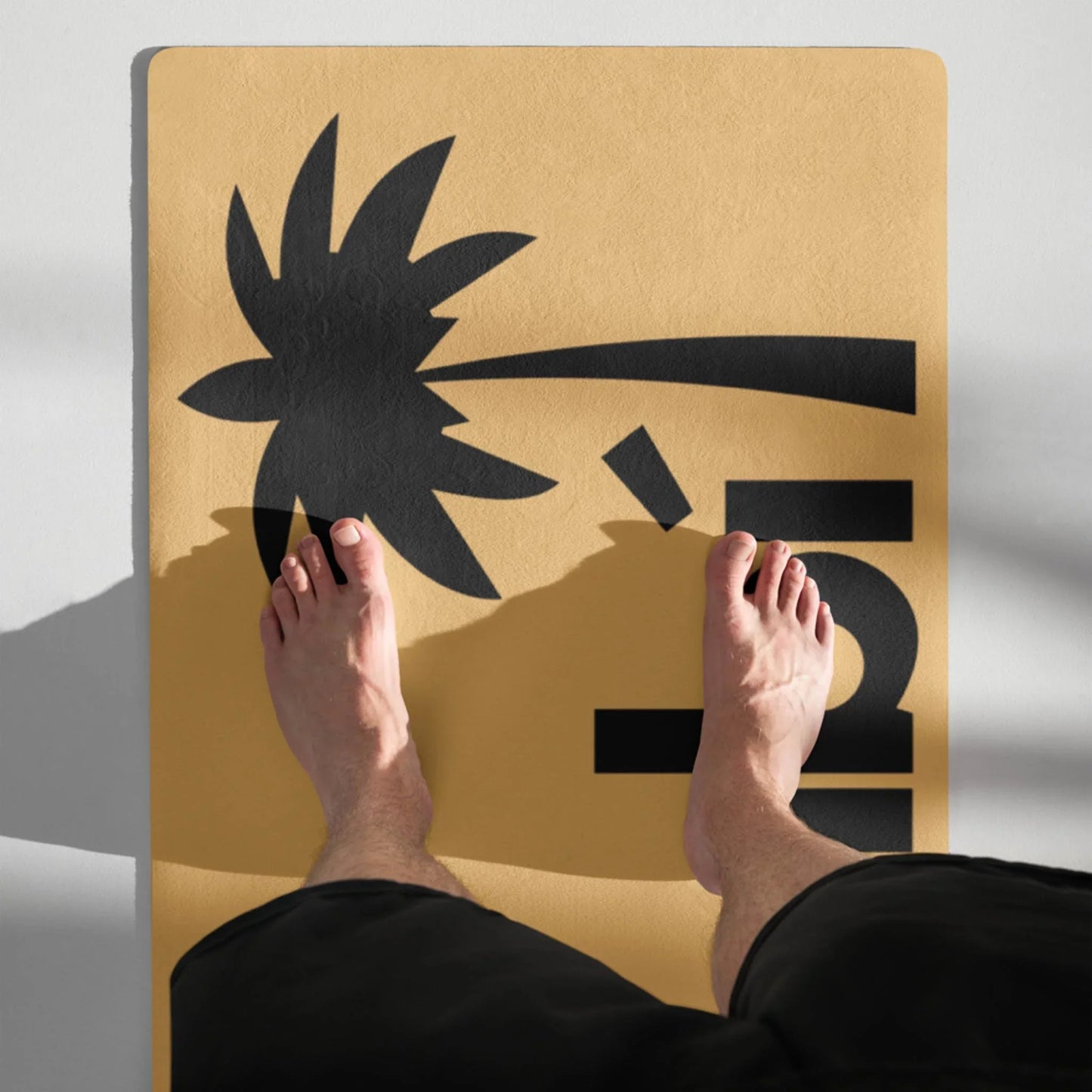 A pair of bare feet stand on a yellow Mambí mat featuring a black graphic of a bent palm tree and the number 5. The persons legs are partially visible, and shadows fall diagonally across the mat.