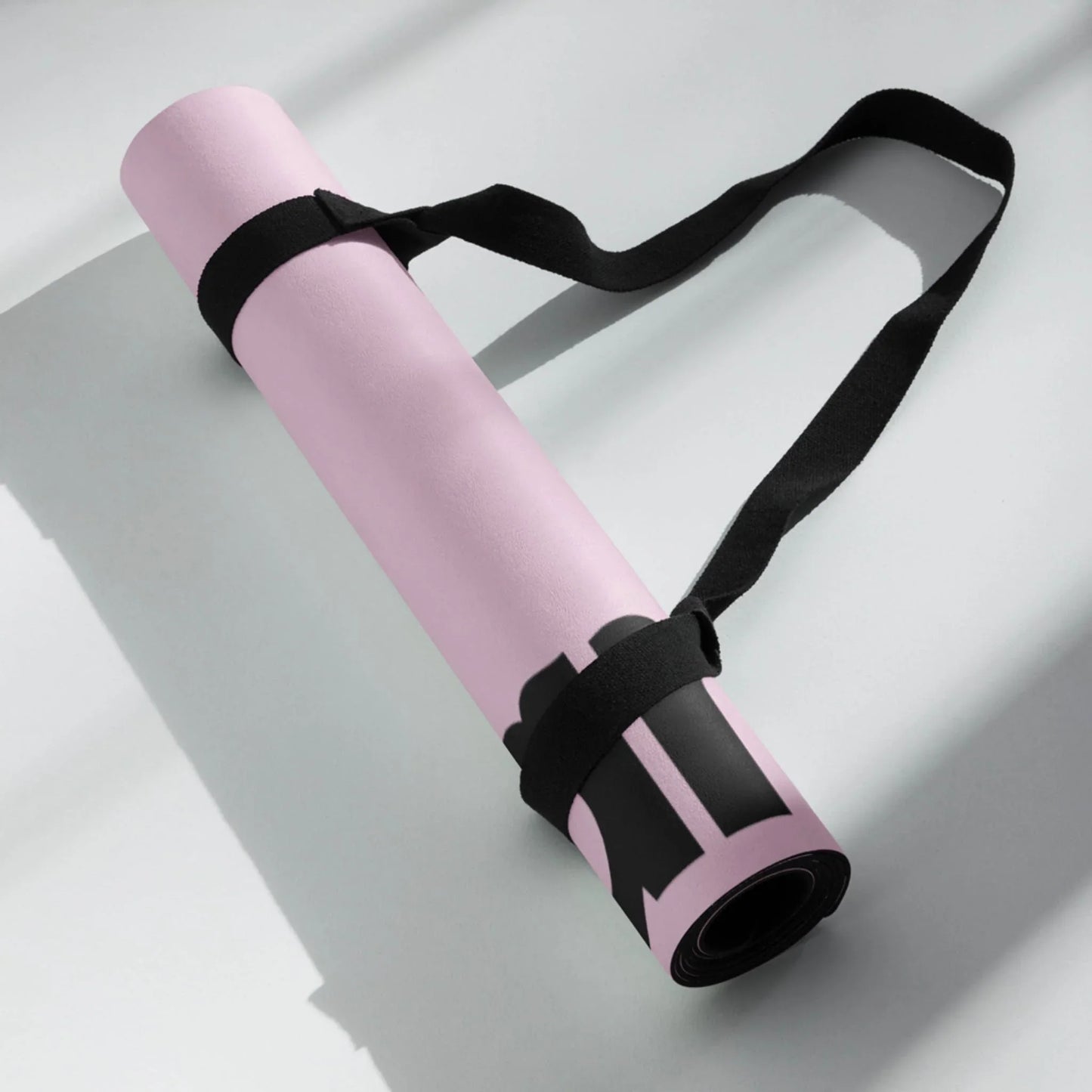 A rolled-up light pink Mambí yoga mat with black straps and a carrying handle rests on a light surface, casting a shadow in the sunlight.