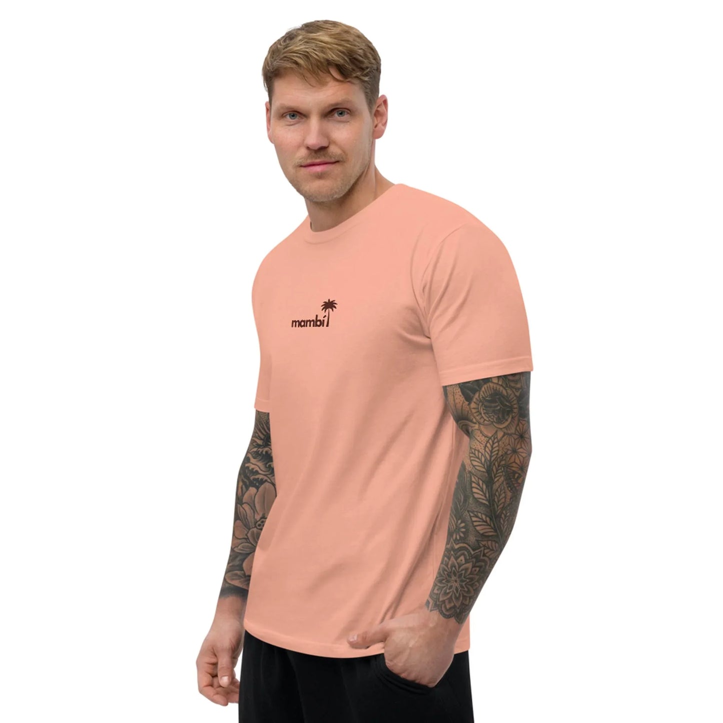 A tattooed man stands sideways with one hand in his pocket, wearing the Printful No Me Jodas Fitted Graphic Tee for men—a soft cotton peach shirt featuring mambi and a small palm tree graphic on the chest.