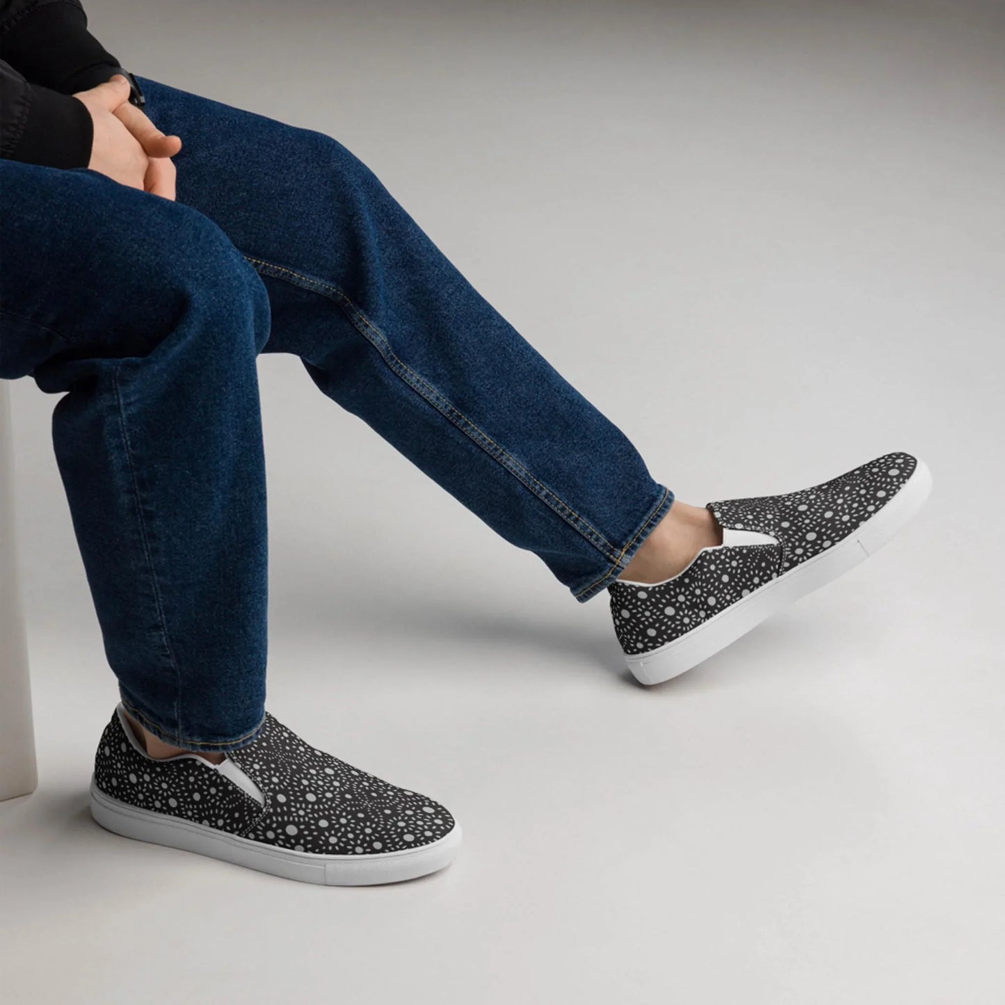 A person wearing blue jeans and black Mambí slip-on shoes with a white speckled pattern sits on a white bench with legs crossed against a plain, light background.