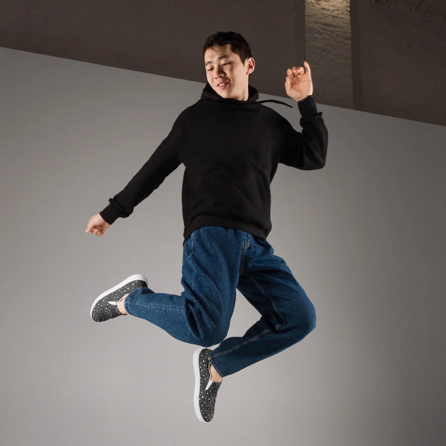 A person wearing a black hoodie, blue jeans, and patterned sneakers is mid-jump against a plain gray background, capturing a joyful and energetic Mambí spirit.