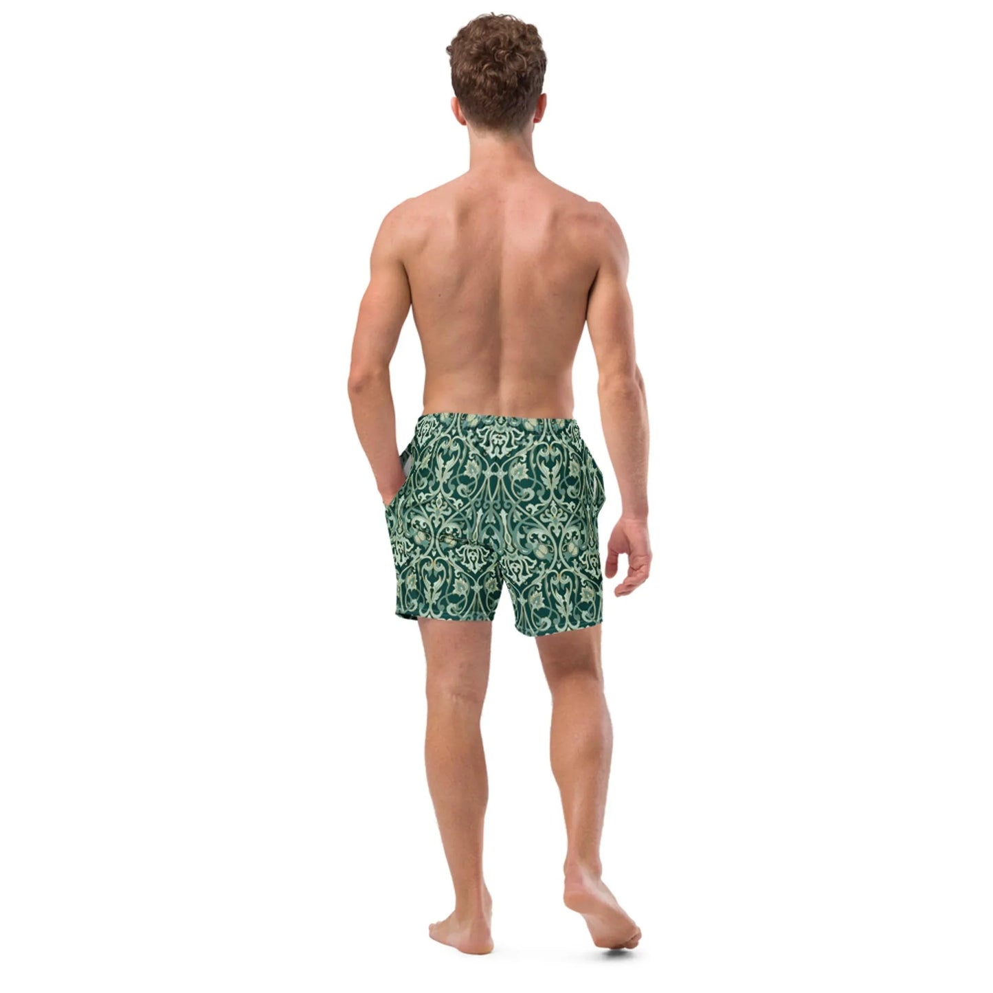 A shirtless man stands barefoot, facing away, with one hand in the pocket of his dark green Oz Swim Trunks by Printful, featuring a light ornate pattern and four-way stretch, water-repellent fabric.