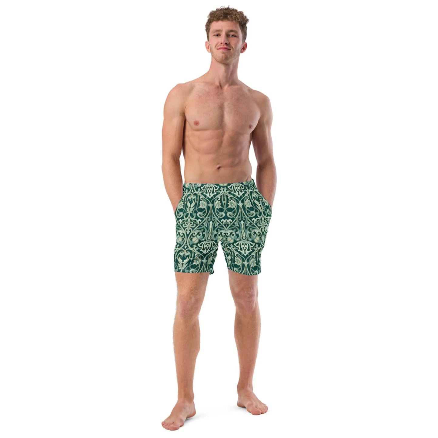 A barefoot man stands confidently with hands in pockets, shirtless and looking upward, wearing Printfuls Oz Swim Trunks for men—green patterned, four-way stretch—set against a plain white background.