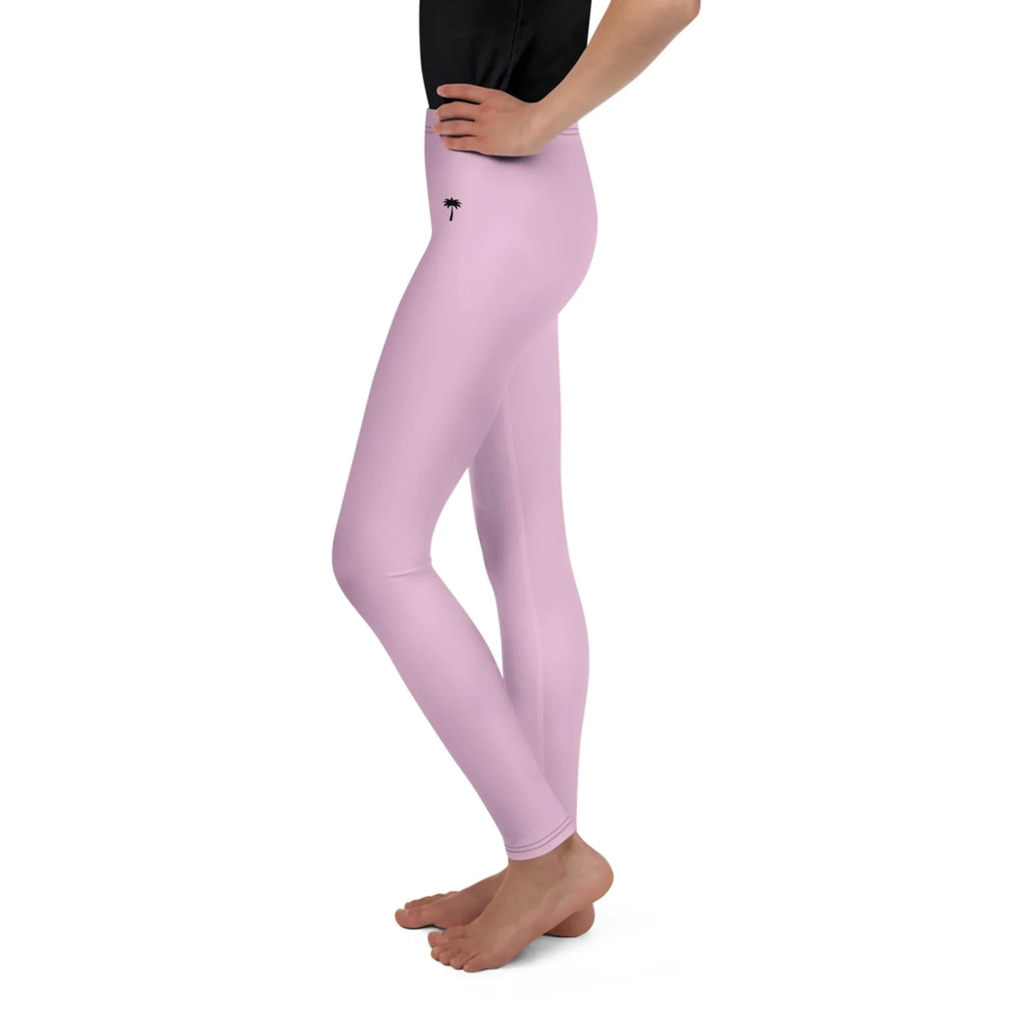 A person stands barefoot in a side view, wearing light pink Mambí leggings with a small palm tree graphic near the left hip and a black top. The background is white.