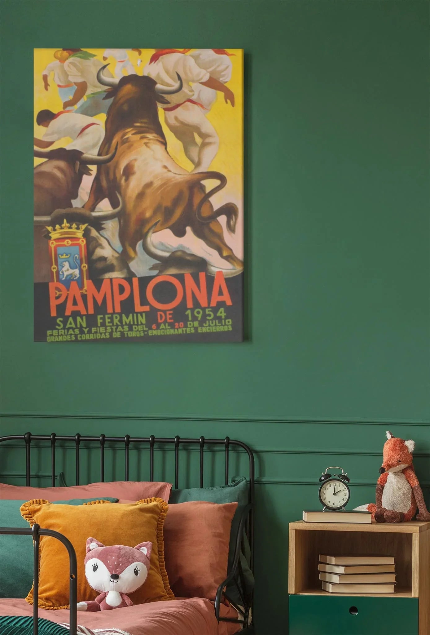 A bedroom with a green wall, orange and pink pillows, a stuffed fox toy, and a nightstand is complete with Printfuls Pamplona reprinted vintage poster on premium luster paper (24x36) displayed above the bed.