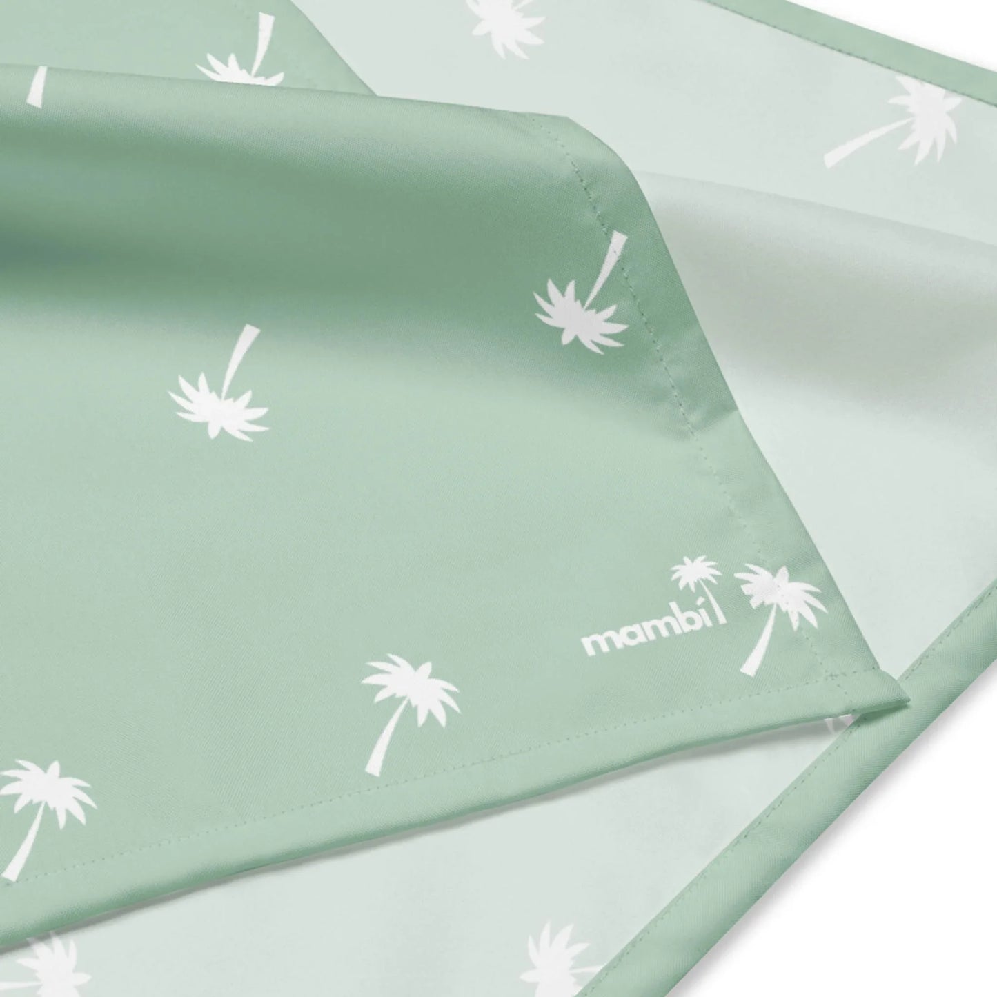 A close-up of light green fabric with white palm tree patterns and the word Mambí in the corner. The soft fabric features stitched edges.