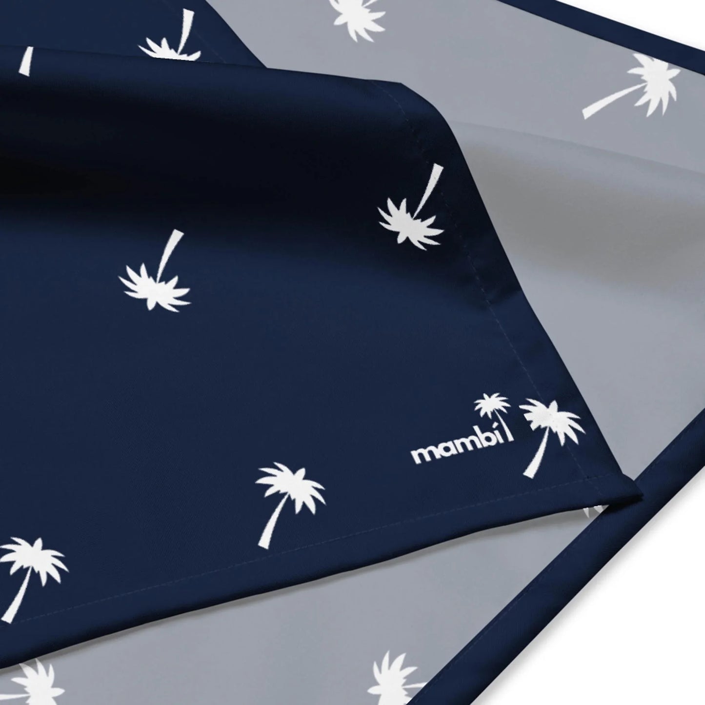 A close-up of a reversible fabric displays a navy side with white palm tree prints and Mambí in the corner, while the underside is solid light gray.
