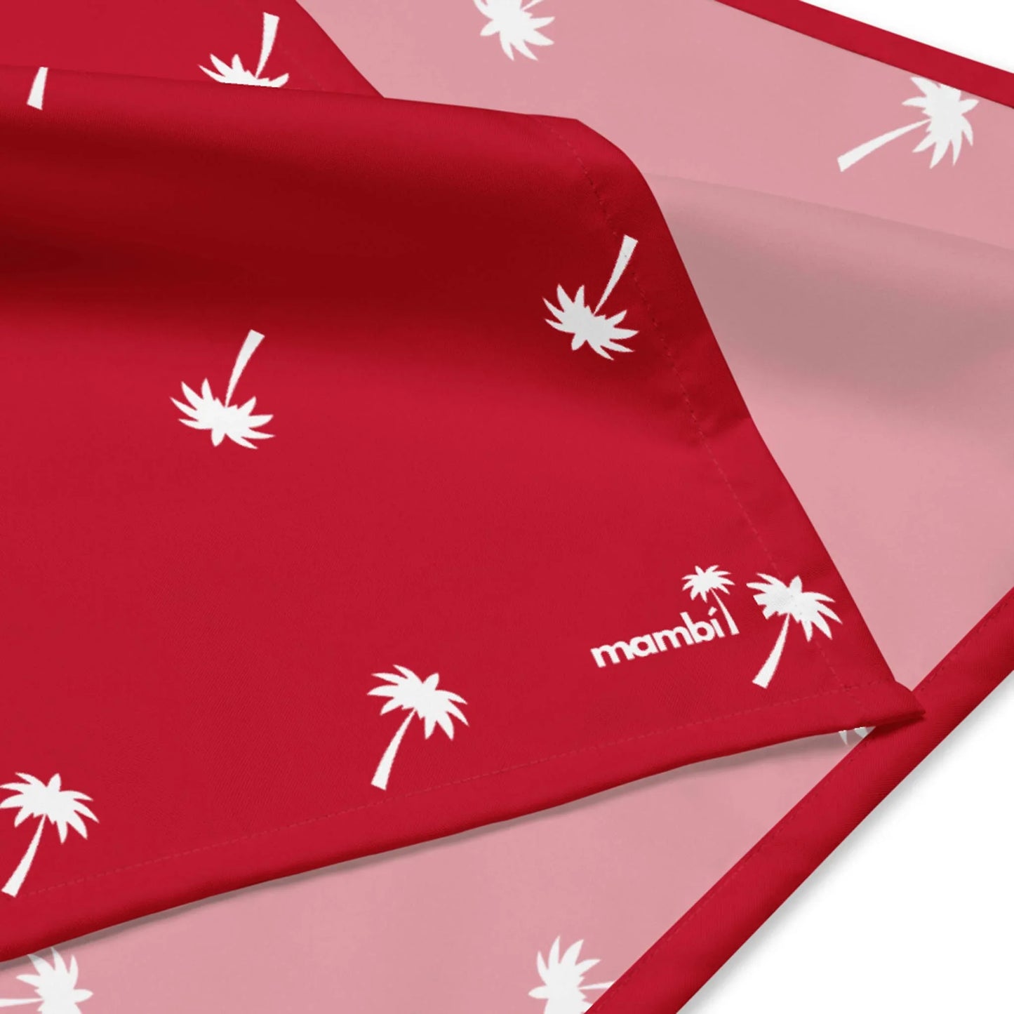 A close-up of red and pink fabric with a white palm tree pattern and Mambí printed in white on the corner. The fabric is partially folded to reveal both sides.