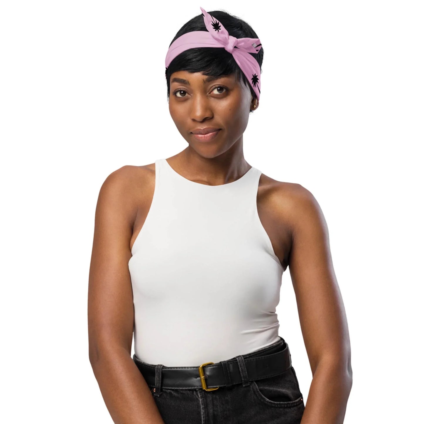 A woman wearing a sleeveless white top, Mambí black high-waisted jeans with a belt, and a pink headband with black designs stands against a white background, smiling softly.