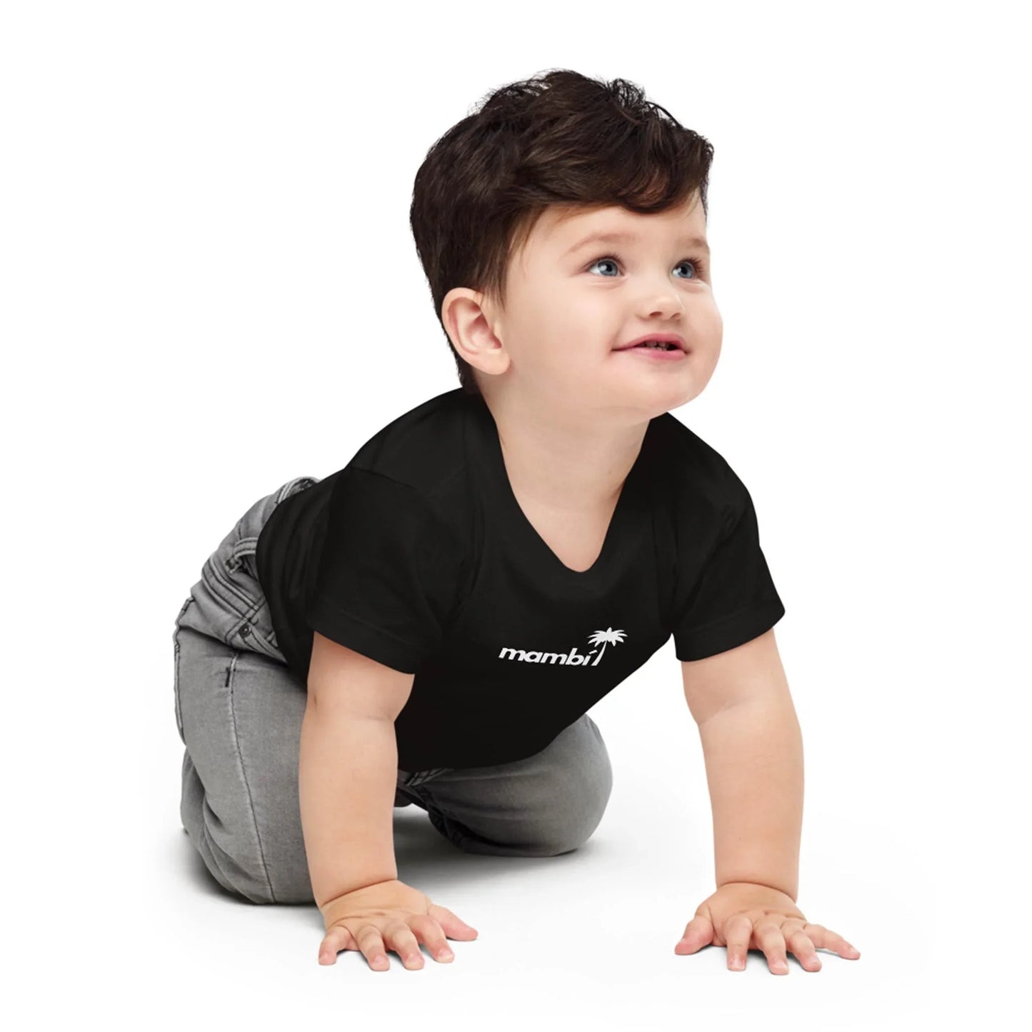 A toddler with dark hair, wearing a black t-shirt and gray pants, is smiling and crawling on the floor. The t-shirt features a small white palm tree logo with the word Mambí on it.