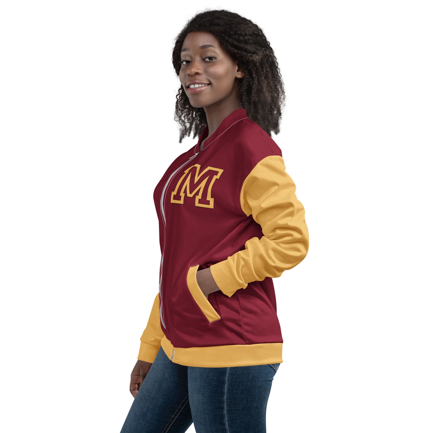 A woman wearing a maroon and gold varsity bomber jacket with premium stitching and a large yellow M on the chest stands with her hands in her pockets, smiling and facing sideways against a white background.