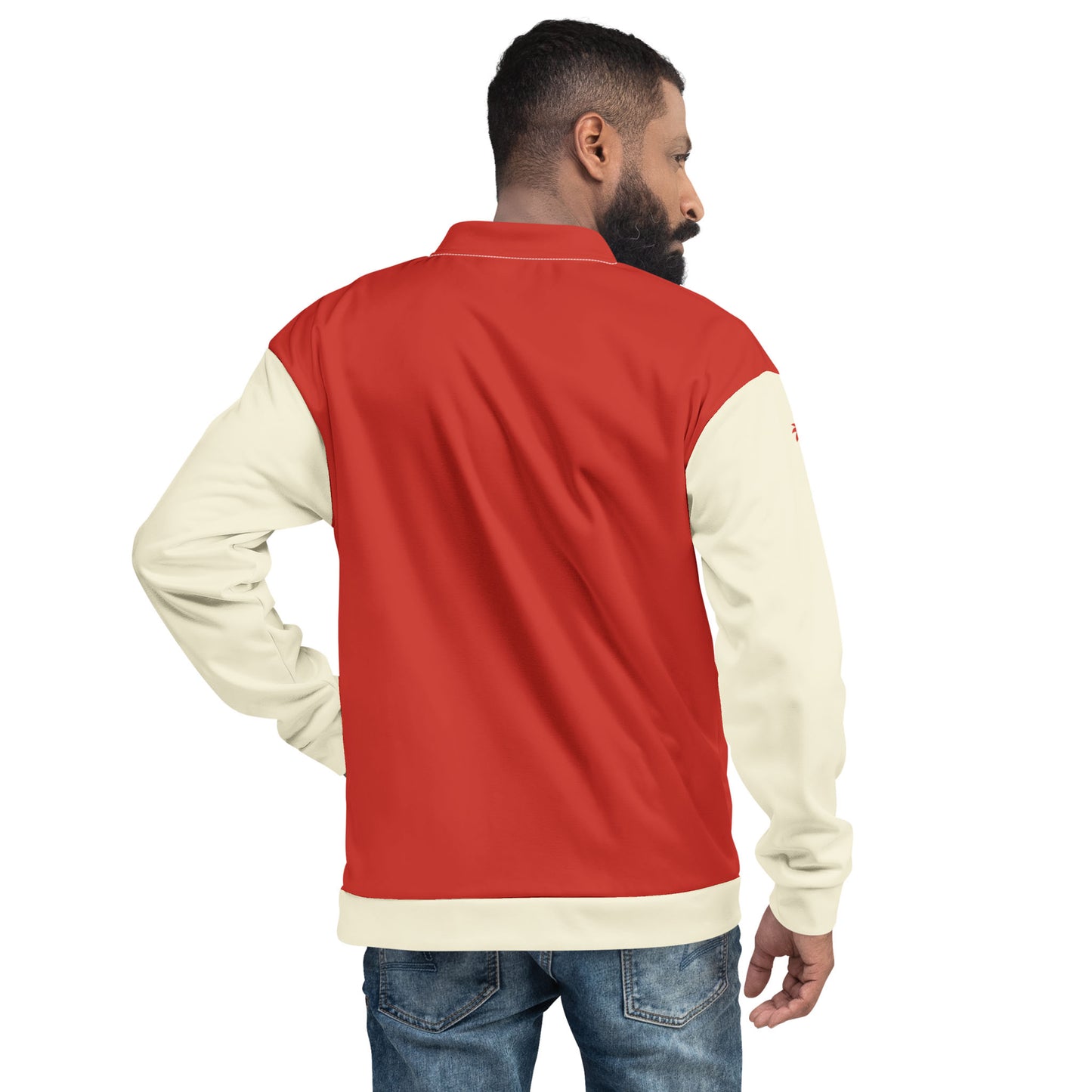 A man stands with his back to the camera, wearing a red and cream varsity bomber jacket with premium stitching and blue jeans. He has a trimmed beard and short hair, and his left hand rests on his hip.