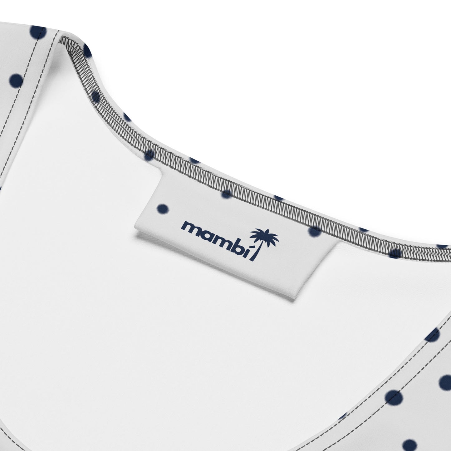 Close-up of the inside neckline of an all-over printed dress with white fabric, navy blue polka dots, and gray stitching. A white tag displays the brand name “mambi” with a palm tree graphic in navy blue.