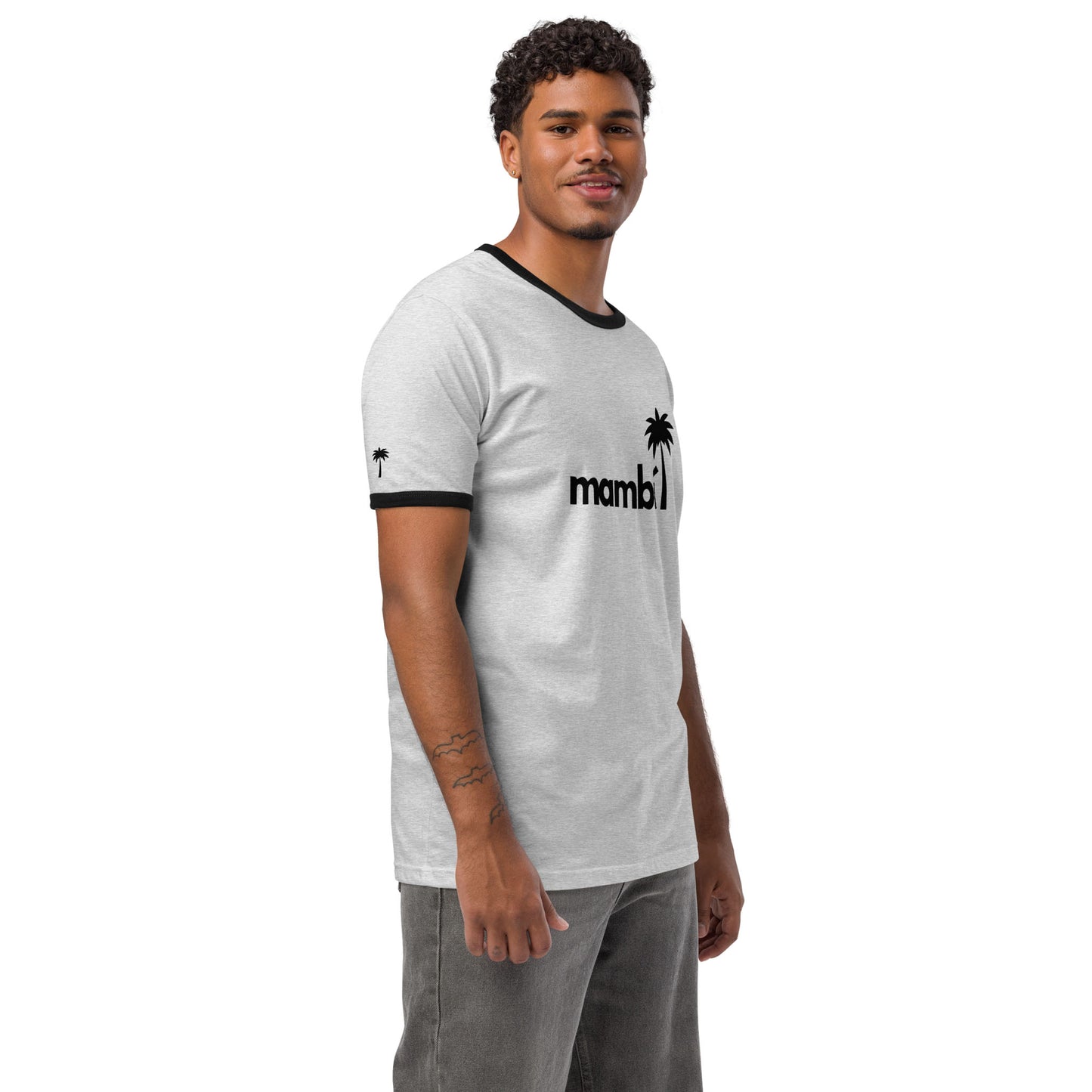 A man with short curly hair and a trimmed beard wears a relaxed fit, lightweight cotton tee with black collar and sleeve hems. The shirt features mambi with a palm tree graphic, giving it a retro look. He stands smiling against a white background.