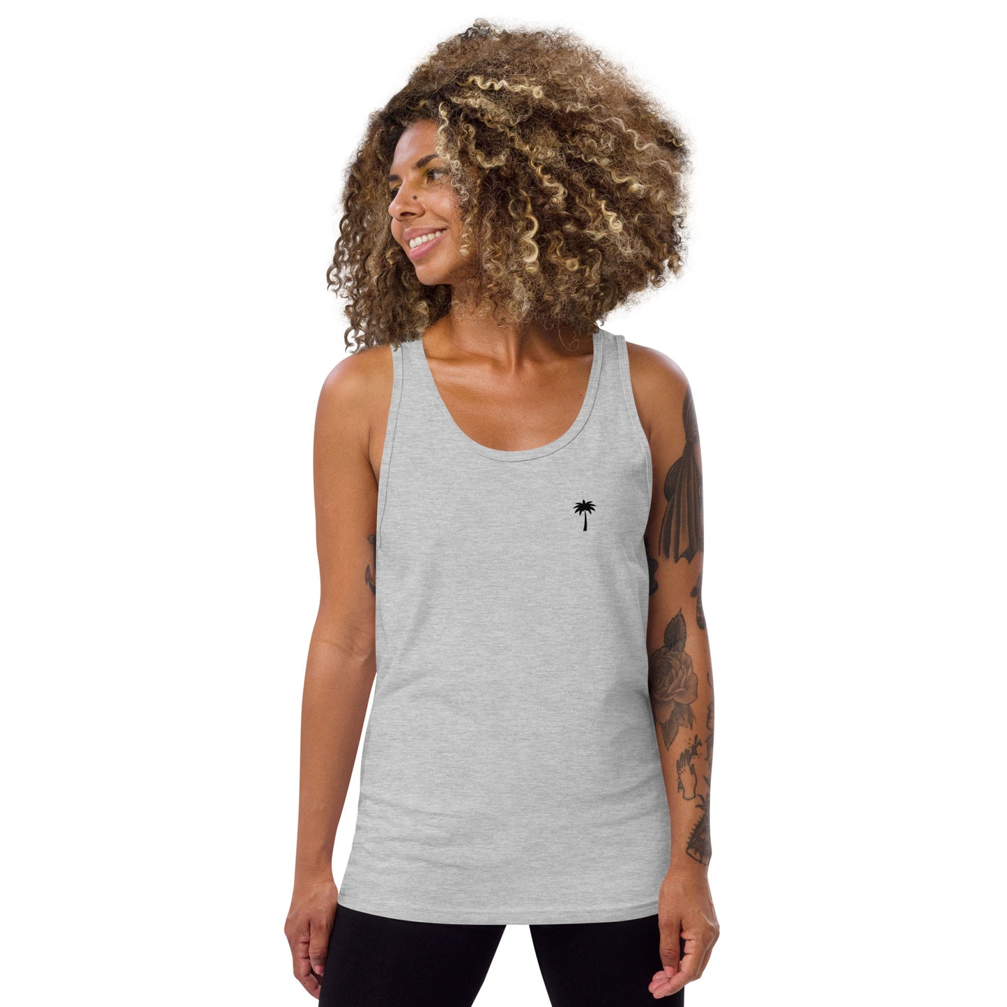 A smiling woman with curly hair wears a classic style light gray tank top, featuring a small black palm tree graphic on the left chest, standing against a plain white background. She has visible tattoos on her left arm.
