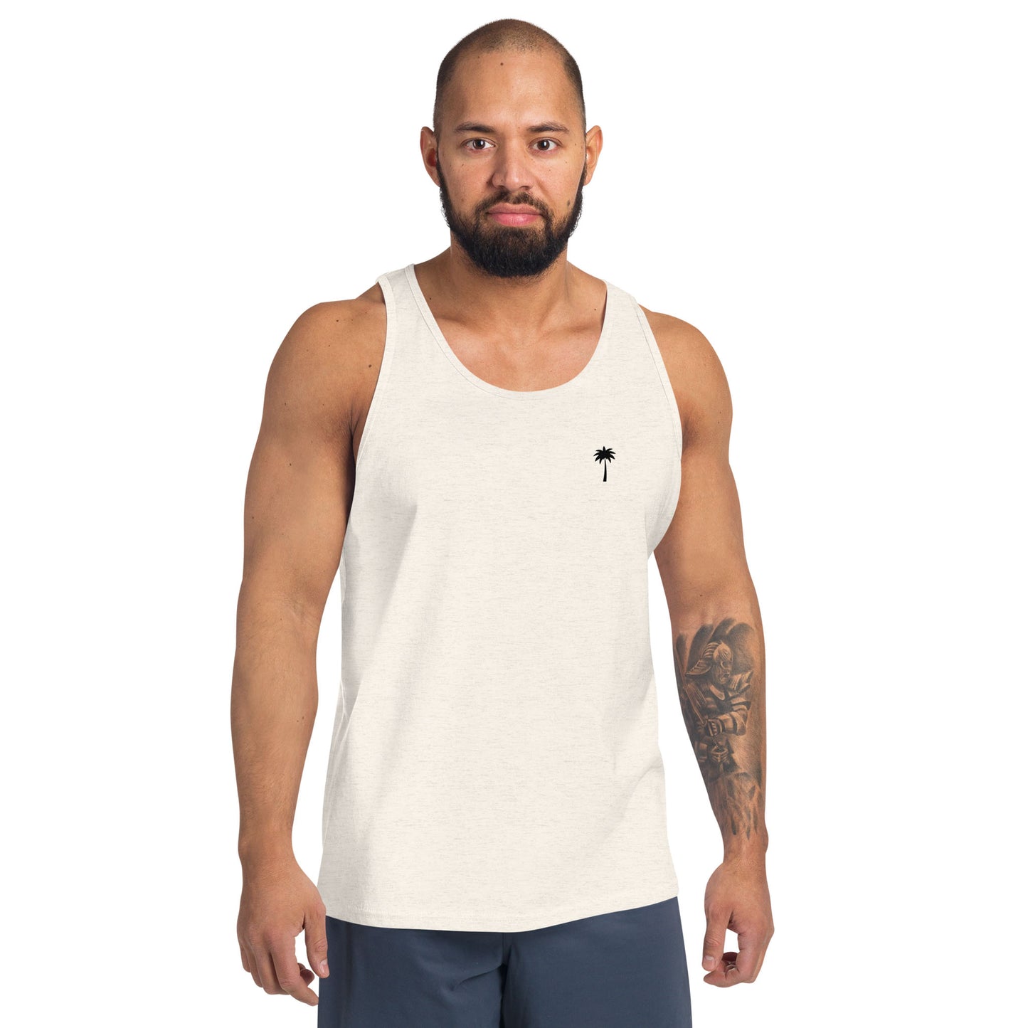 A man with a beard and arm tattoos showcases a classic style in a light-colored tank top made from premium materials, featuring a small palm tree design on the chest and paired with dark shorts against a plain white background.