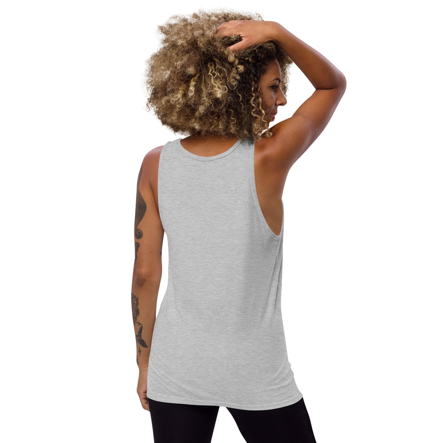 A person with curly hair, tattooed left arm, and wearing a classic style light gray tank top made from premium materials and black pants, stands with their back to the camera and one hand resting on their head.