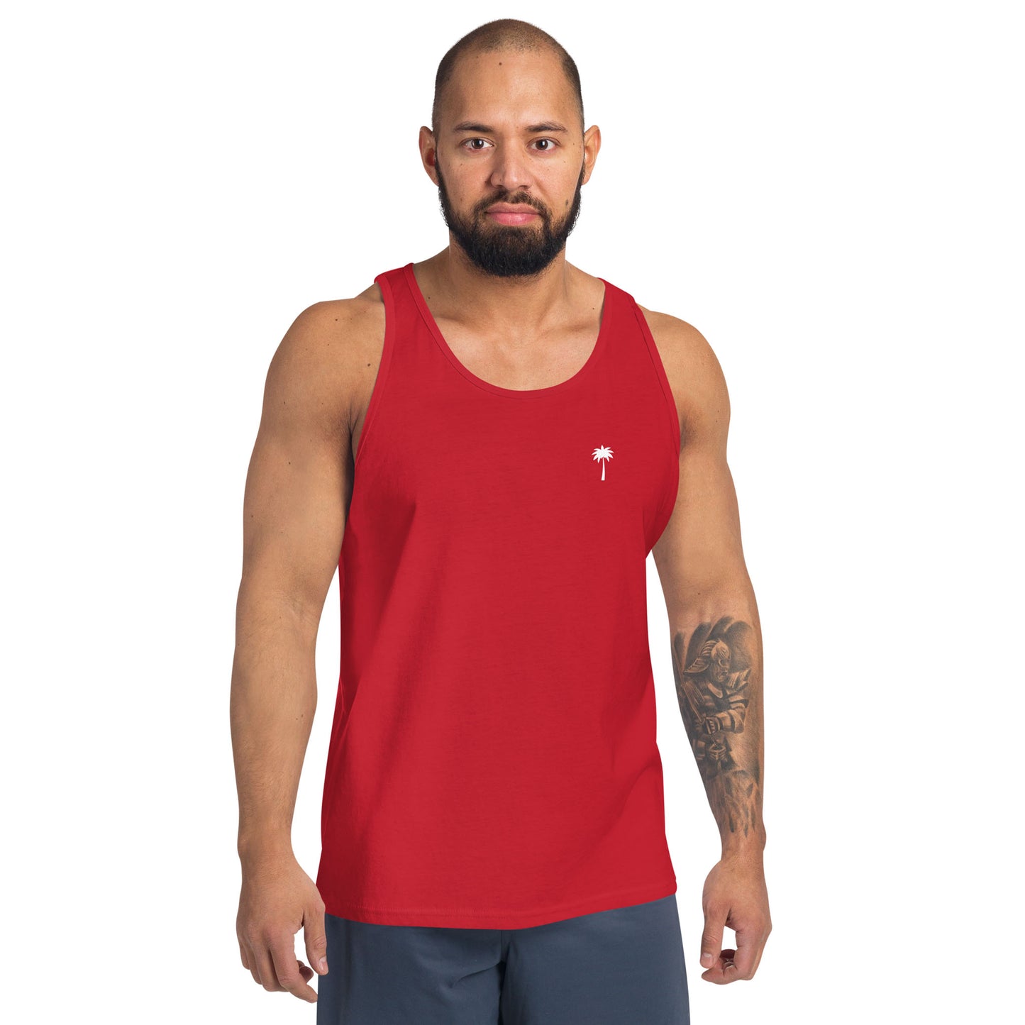 A man with a beard stands facing forward, wearing a classic ringspun cotton tank top with a small white palm tree logo on the chest and dark shorts. He has a tattoo sleeve on his left arm and a neutral expression.