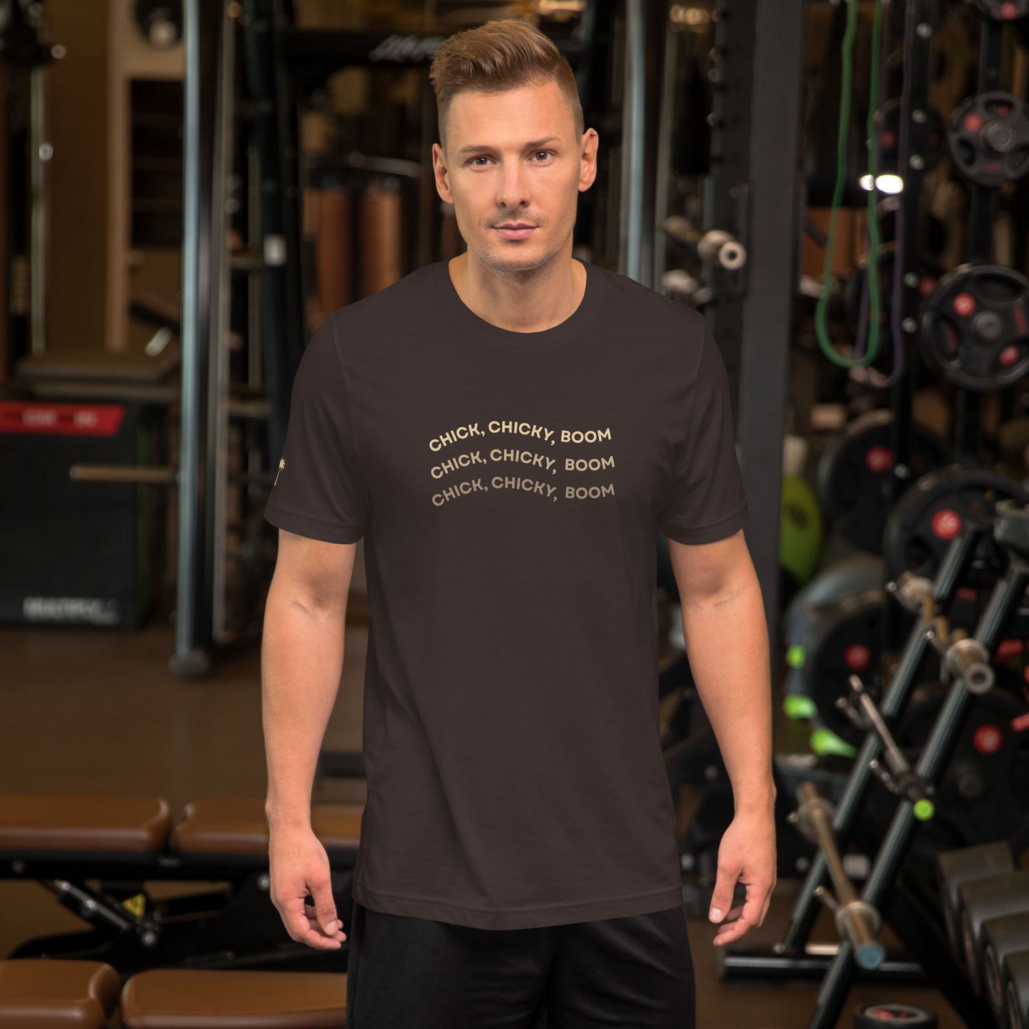 A man stands in a gym wearing a comfortable cotton black fitted shirt with the repeated phrase “CHICK, CHICKY, BOOM” printed on the front in light text. Gym equipment and weights are visible in the background.